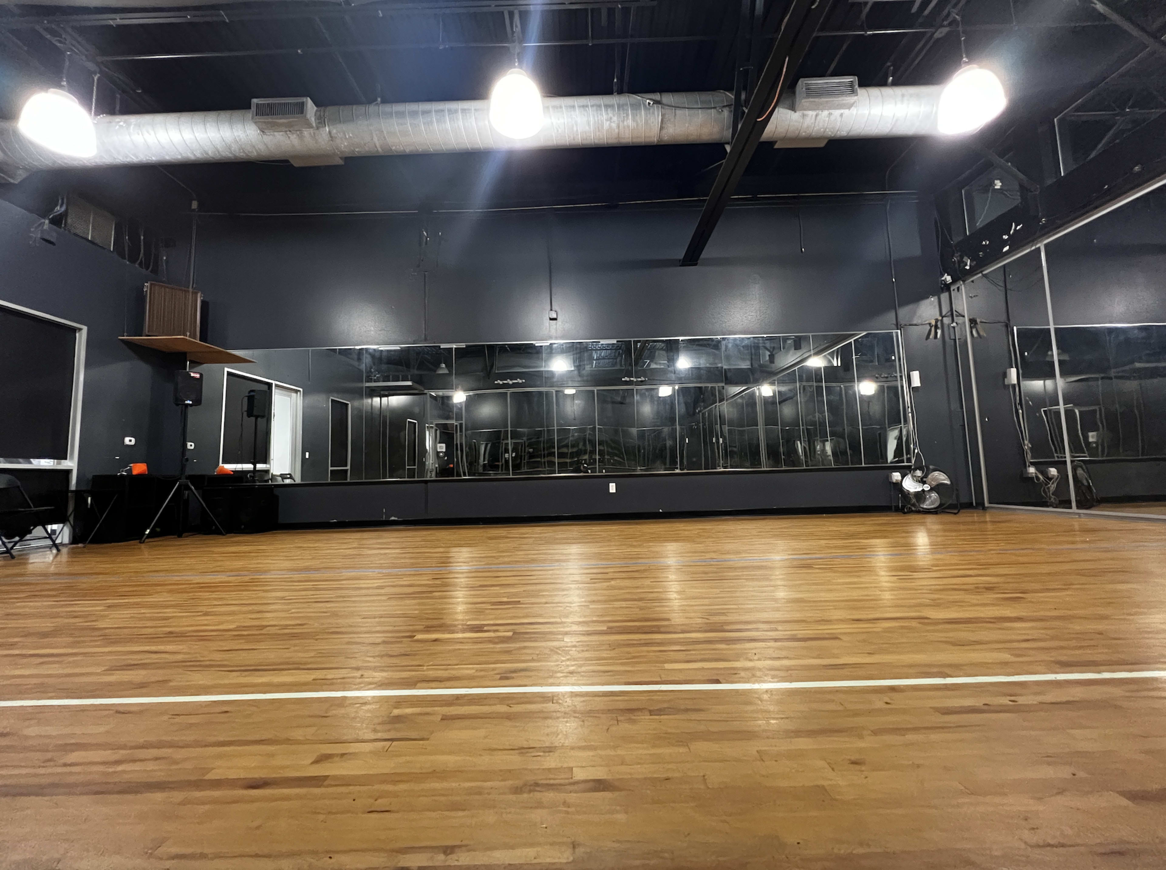 A spacious, empty dance studio with wooden flooring, large mirrors along one wall, and bright overhead lights.