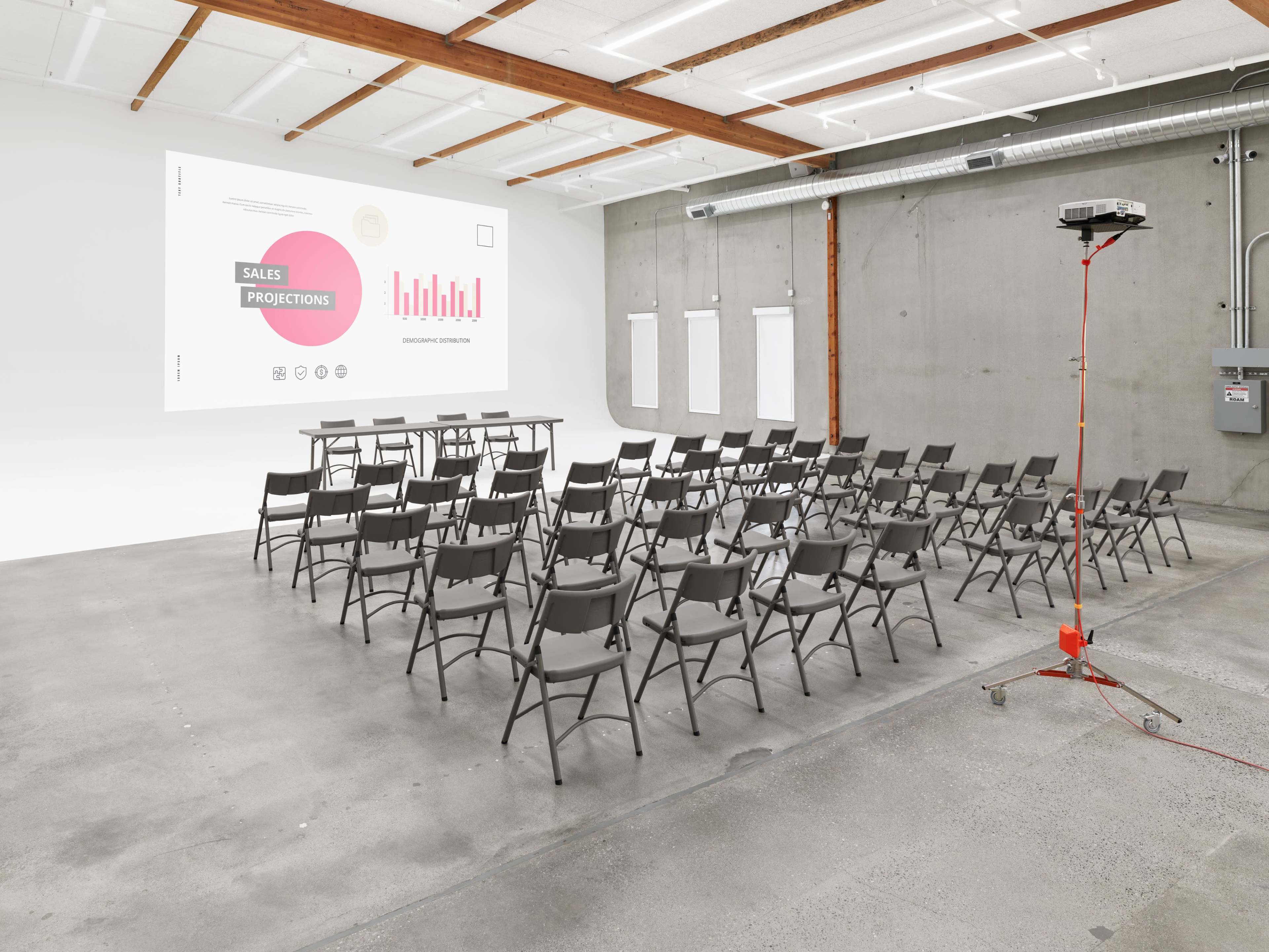 A conference room features rows of gray chairs facing a blank wall with a large projector screen displaying sales projections.