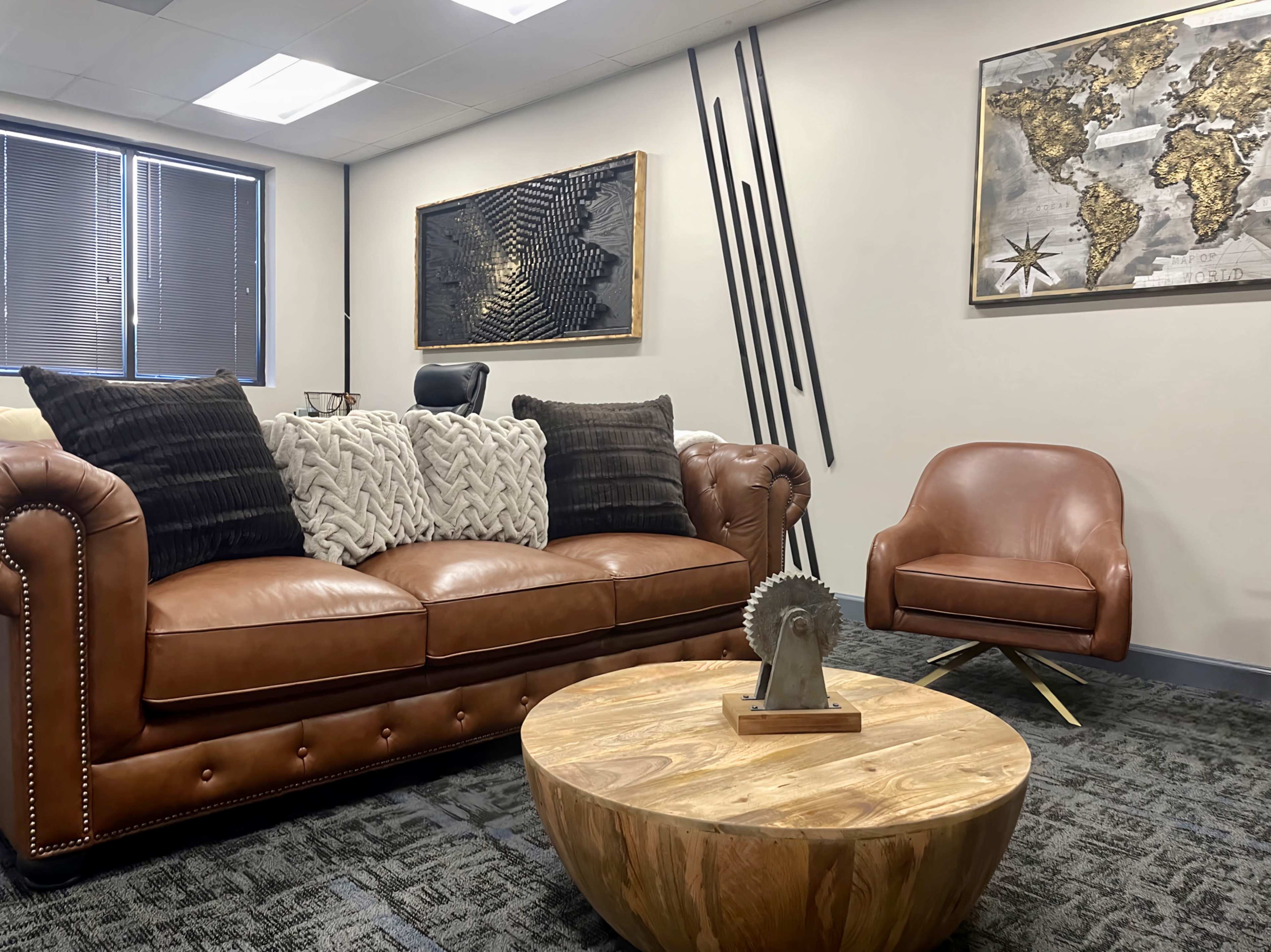 A cozy office space featuring a brown leather sofa with decorative pillows, a wooden coffee table, and a modern armchair, along with wall art including a world map and abstract pieces.