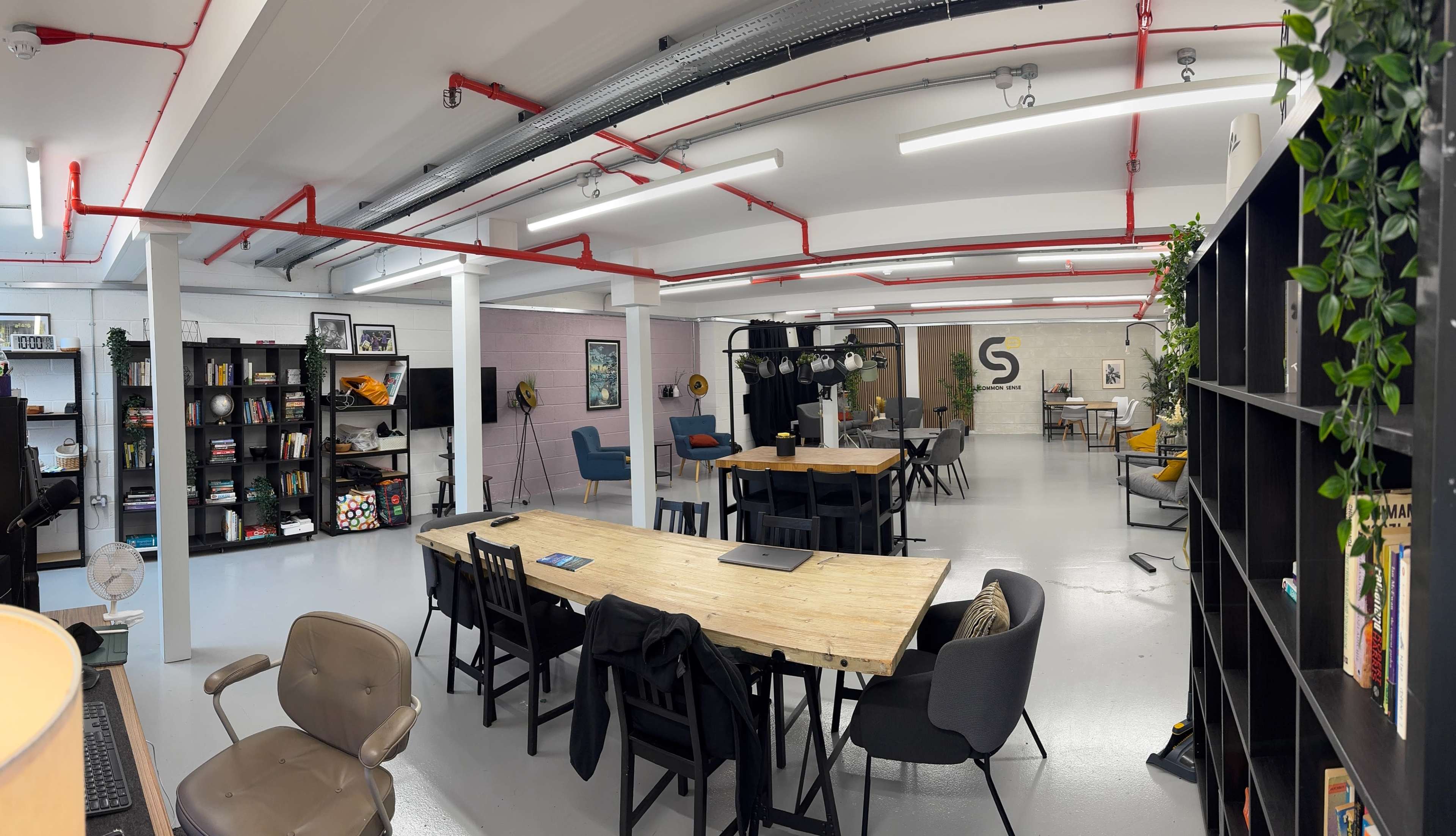 The image shows a modern office space with wooden tables, chairs, shelves filled with books and decorations, and open areas illuminated by overhead lighting.