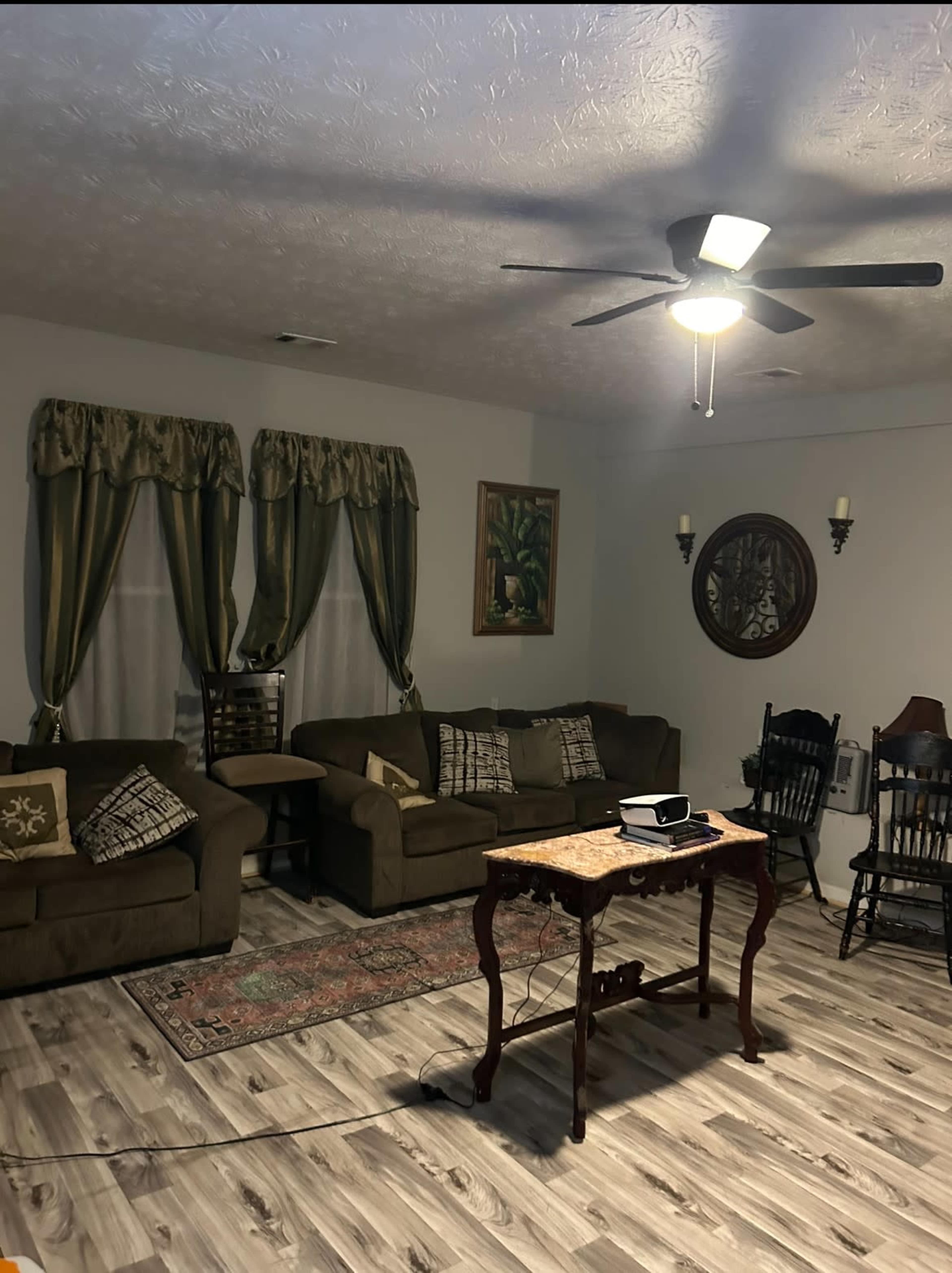 The image shows a living room with two couches, a coffee table, a wooden rocking chair, and decorative curtains, all arranged around a light-colored wooden floor.
