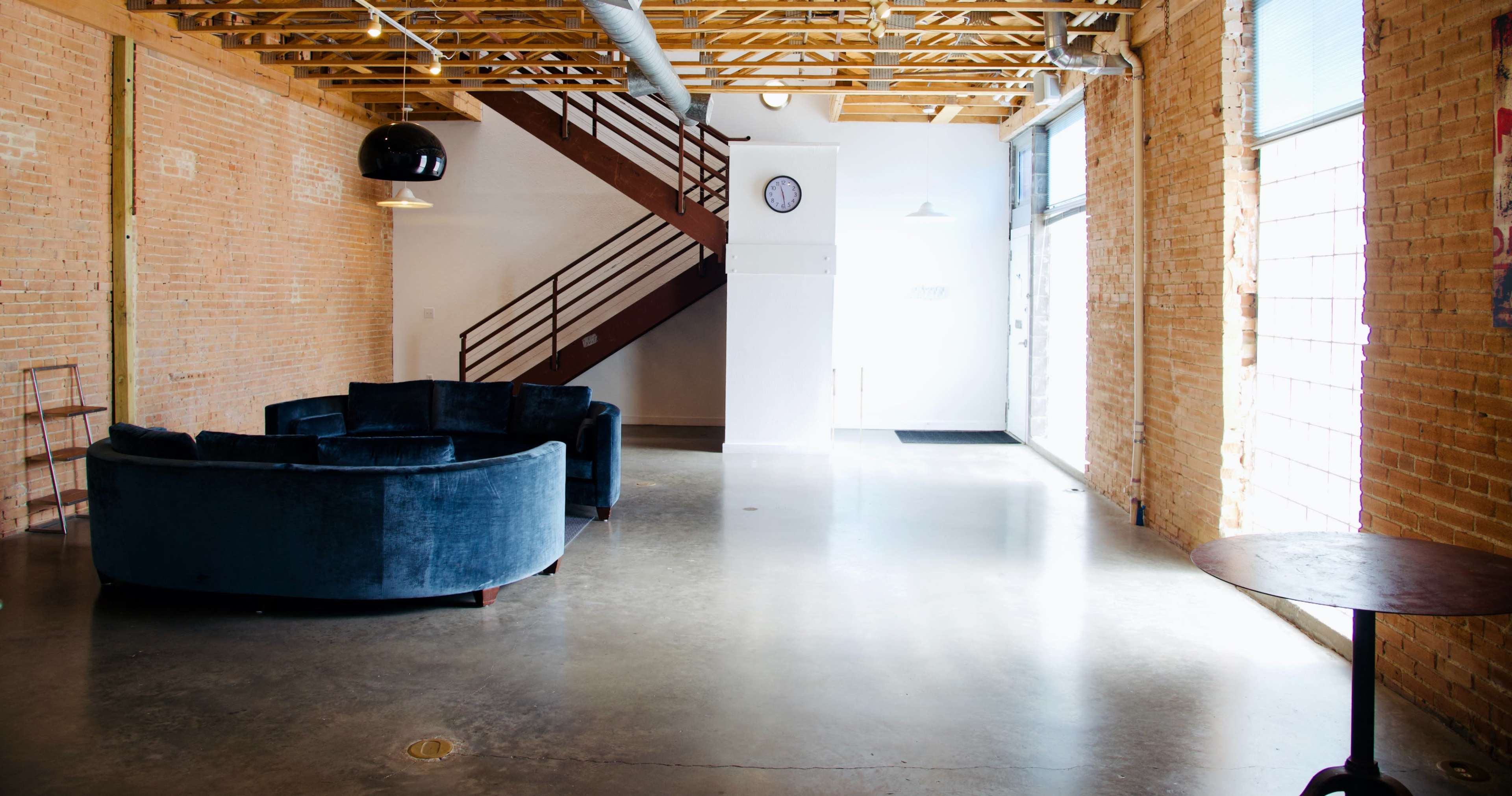 A spacious interior with exposed brick walls, a circular blue velvet couch, a wooden staircase, and large windows allowing light to fill the area.