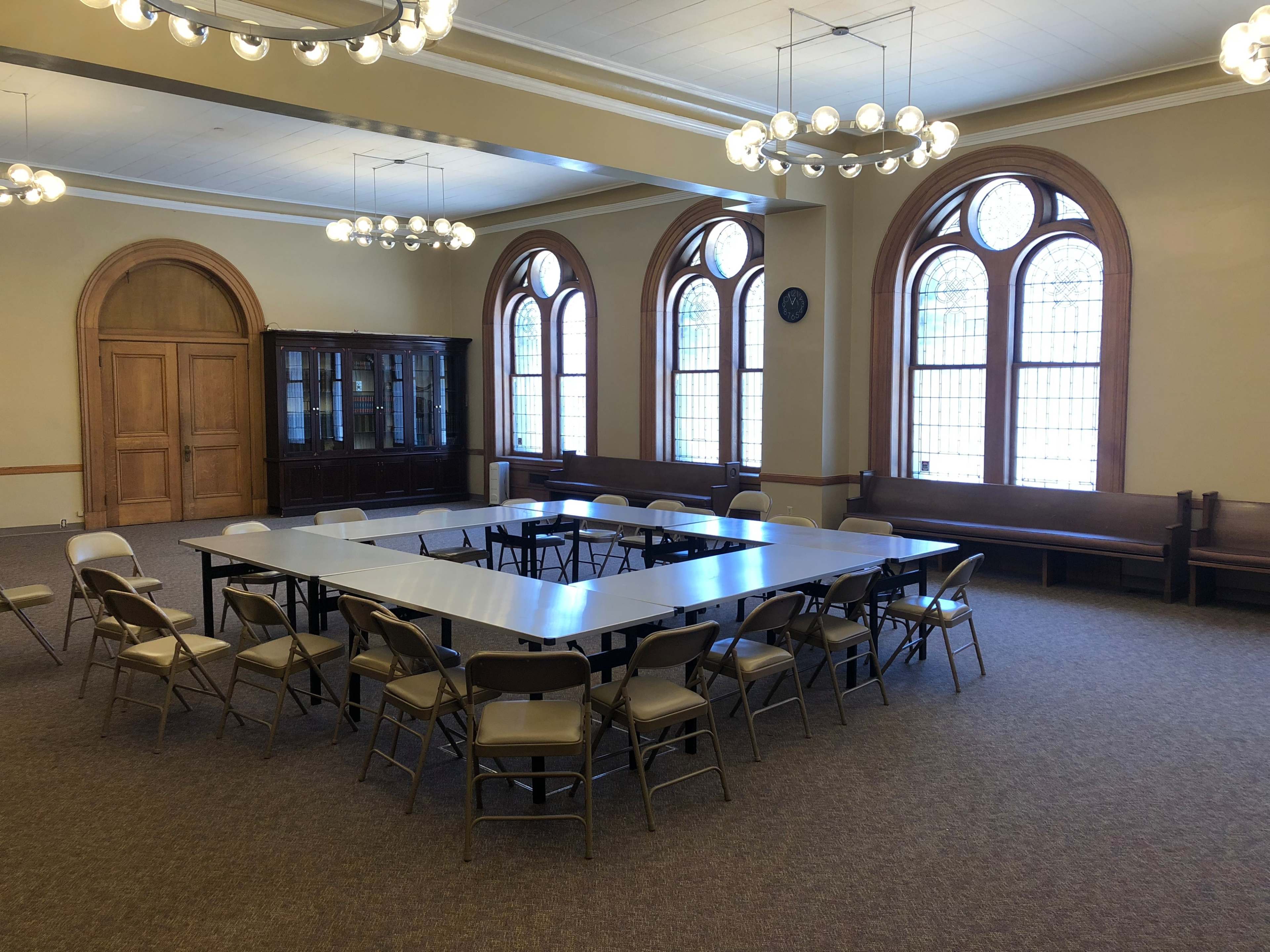 A large meeting room features a rectangular table surrounded by folding chairs, with windows offering natural light and wooden accents.