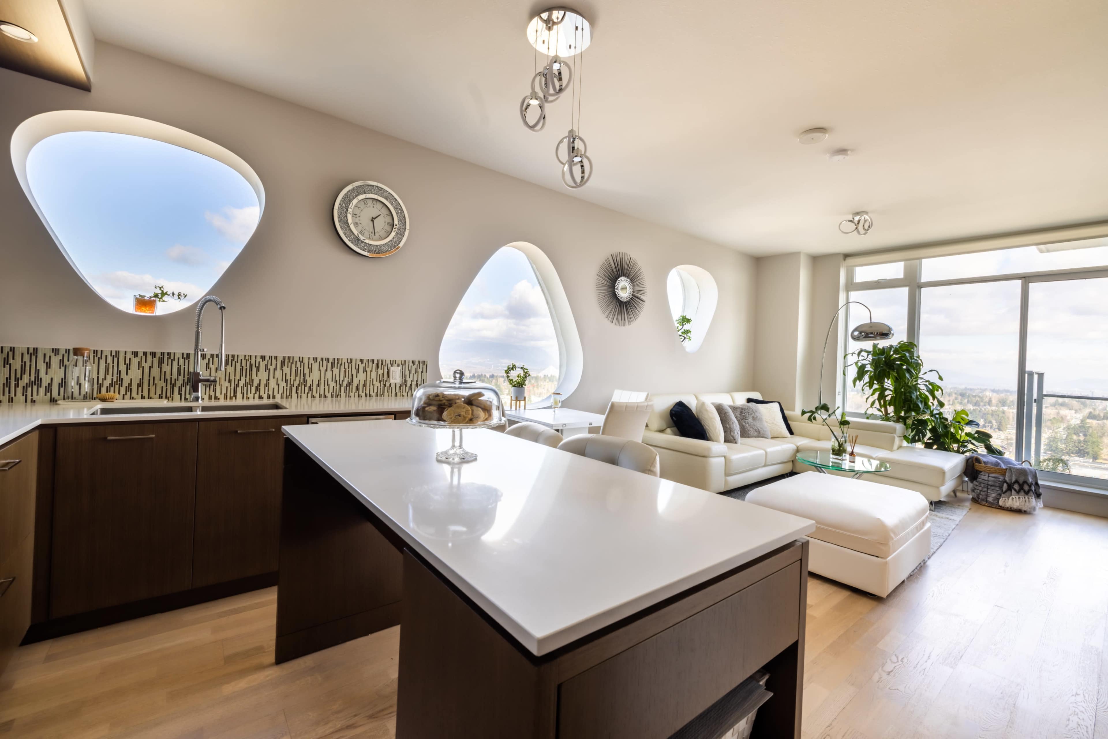 A modern kitchen and living area feature large, uniquely shaped windows, sleek cabinetry, and contemporary furnishings.