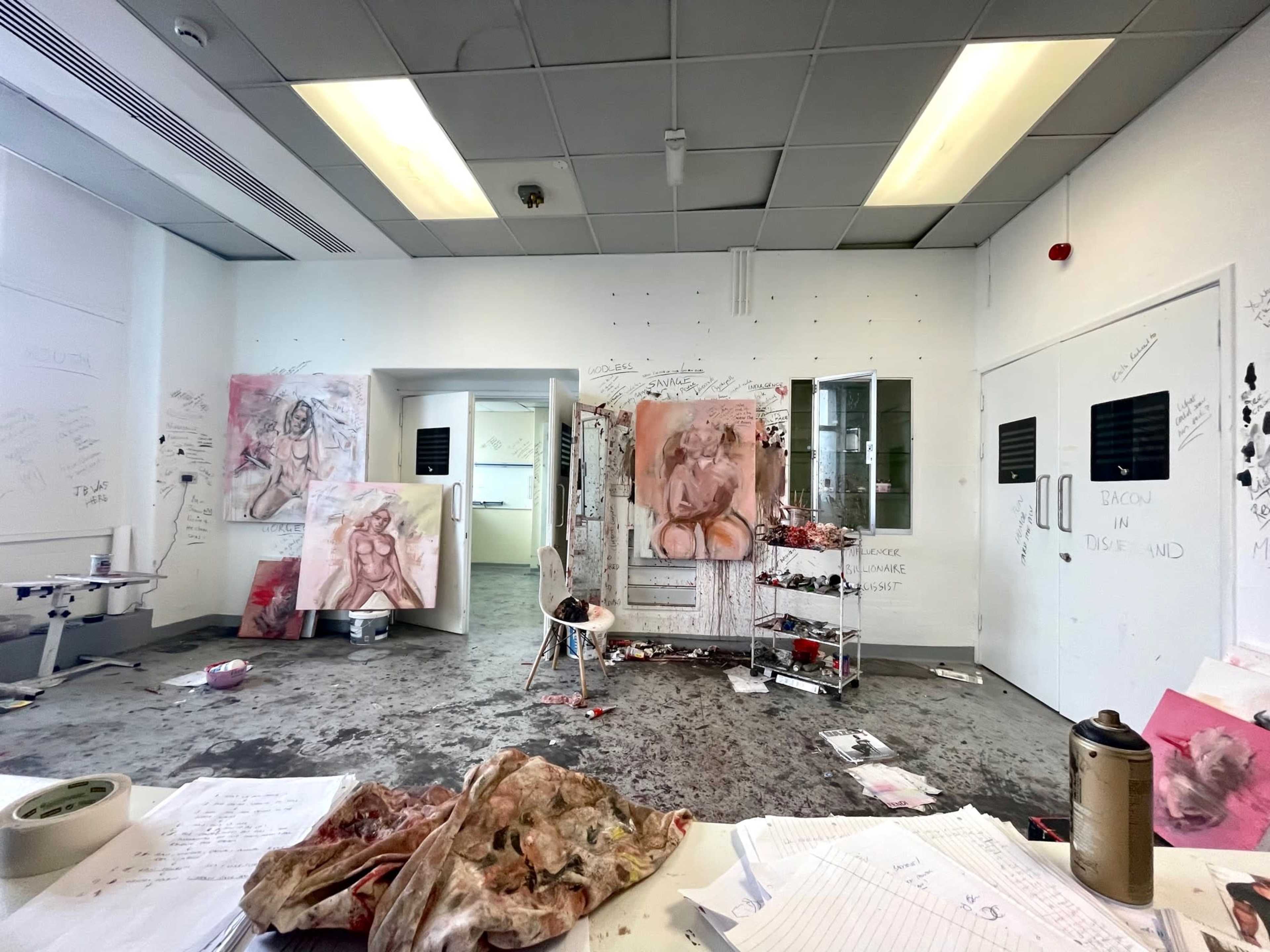 A cluttered art studio with splatters on the floor, unfinished artworks on the walls, and scattered materials on a worktable.