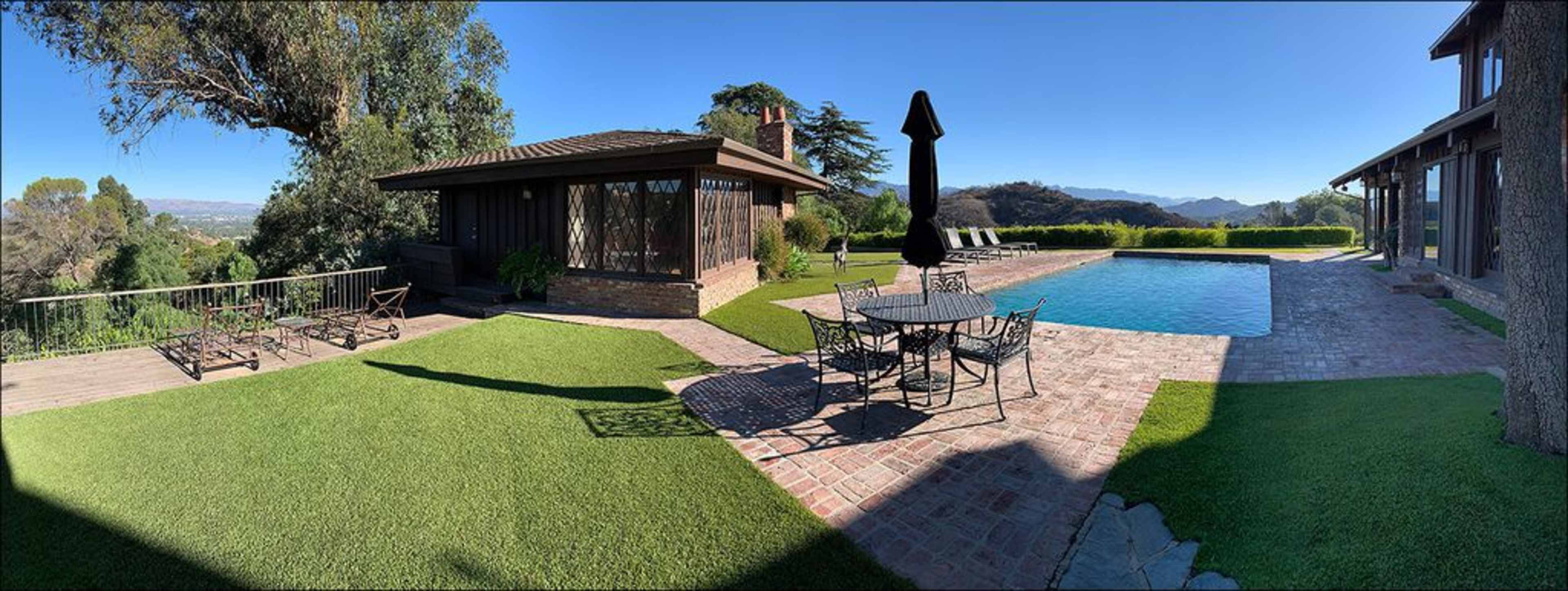 A panoramic view of a landscaped backyard featuring a swimming pool, a stone patio with a dining table, and a separate structure in a hilly area.