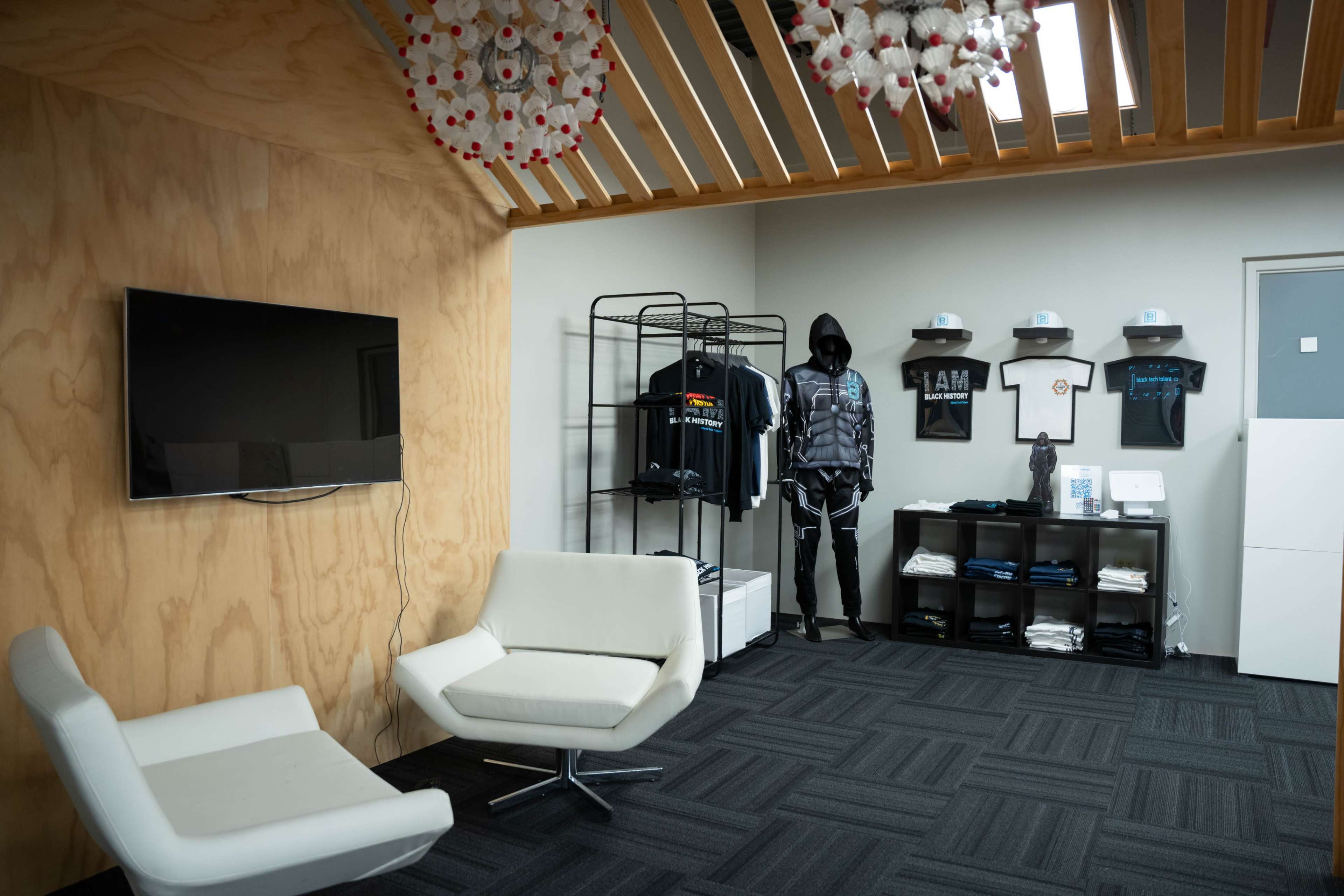 The image shows a modern room with a wooden accent wall, featuring a television, a clothing display with T-shirts and a mannequin dressed in dark attire, and seating in the form of a white chair.