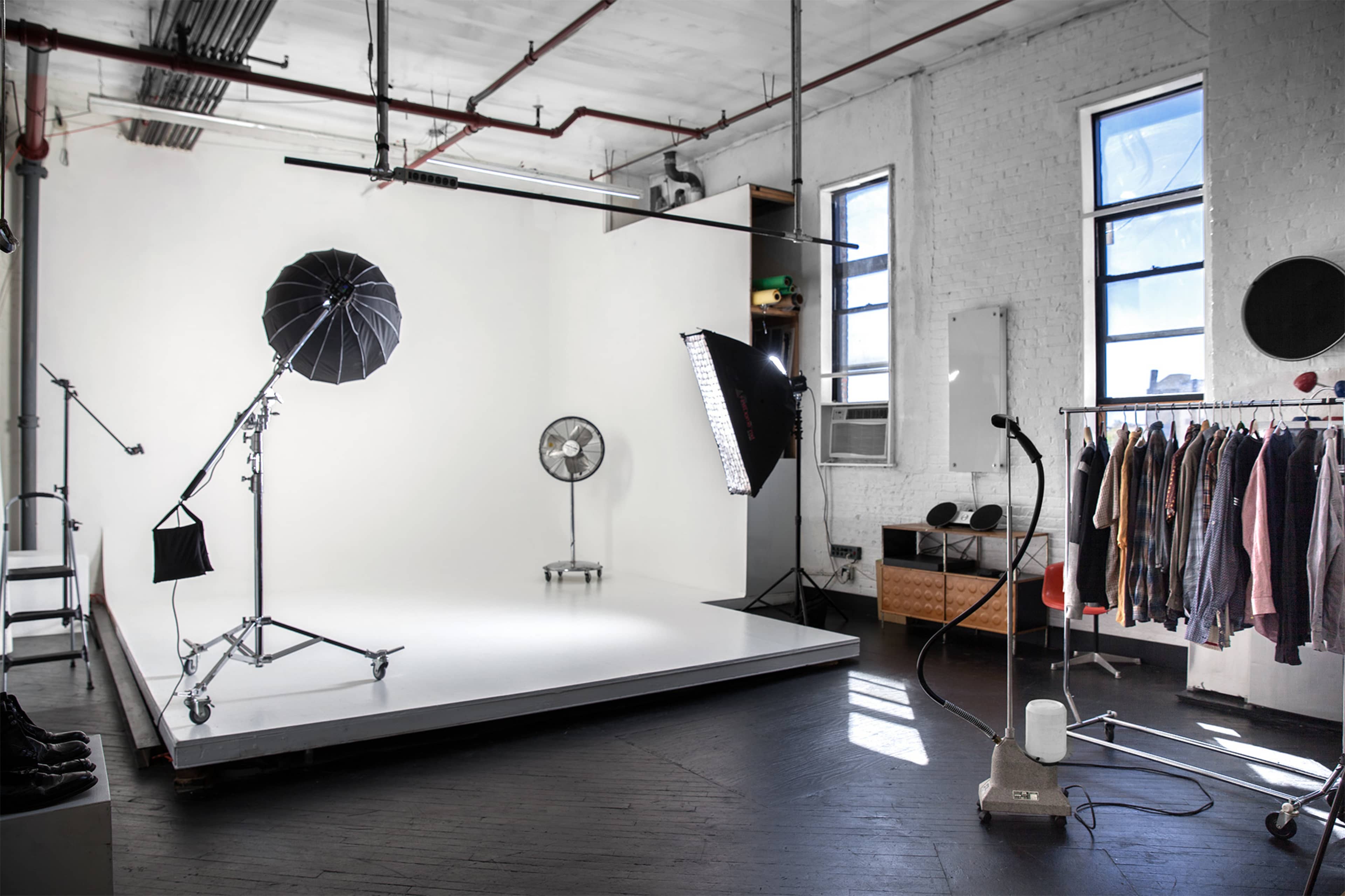 A photography studio features a white backdrop, lighting equipment, a fan, and a clothing rack with various garments.
