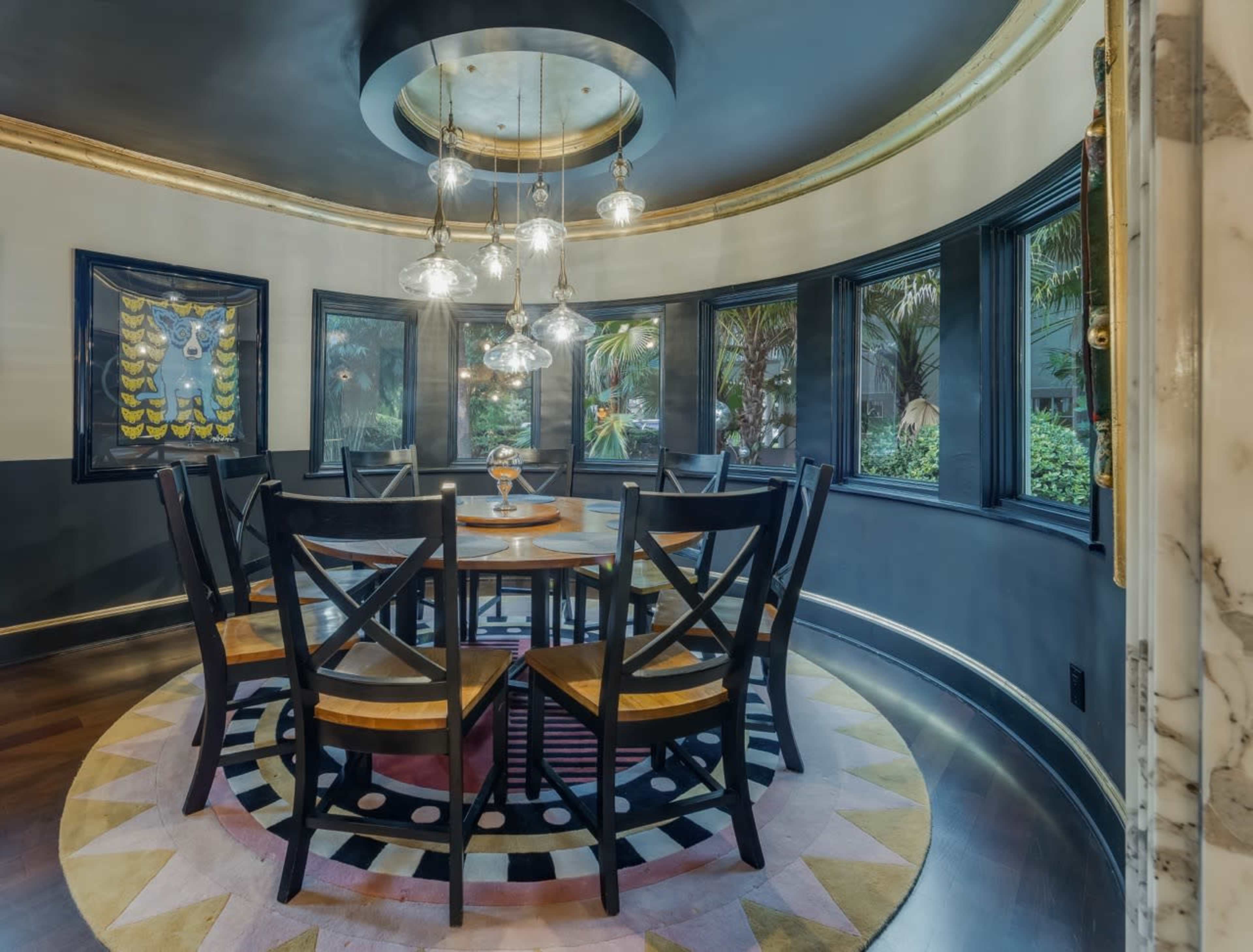 The image shows a circular dining room with a large table surrounded by black wooden chairs, illuminated by multiple pendant lights, and featuring windows that offer a view of greenery outside.