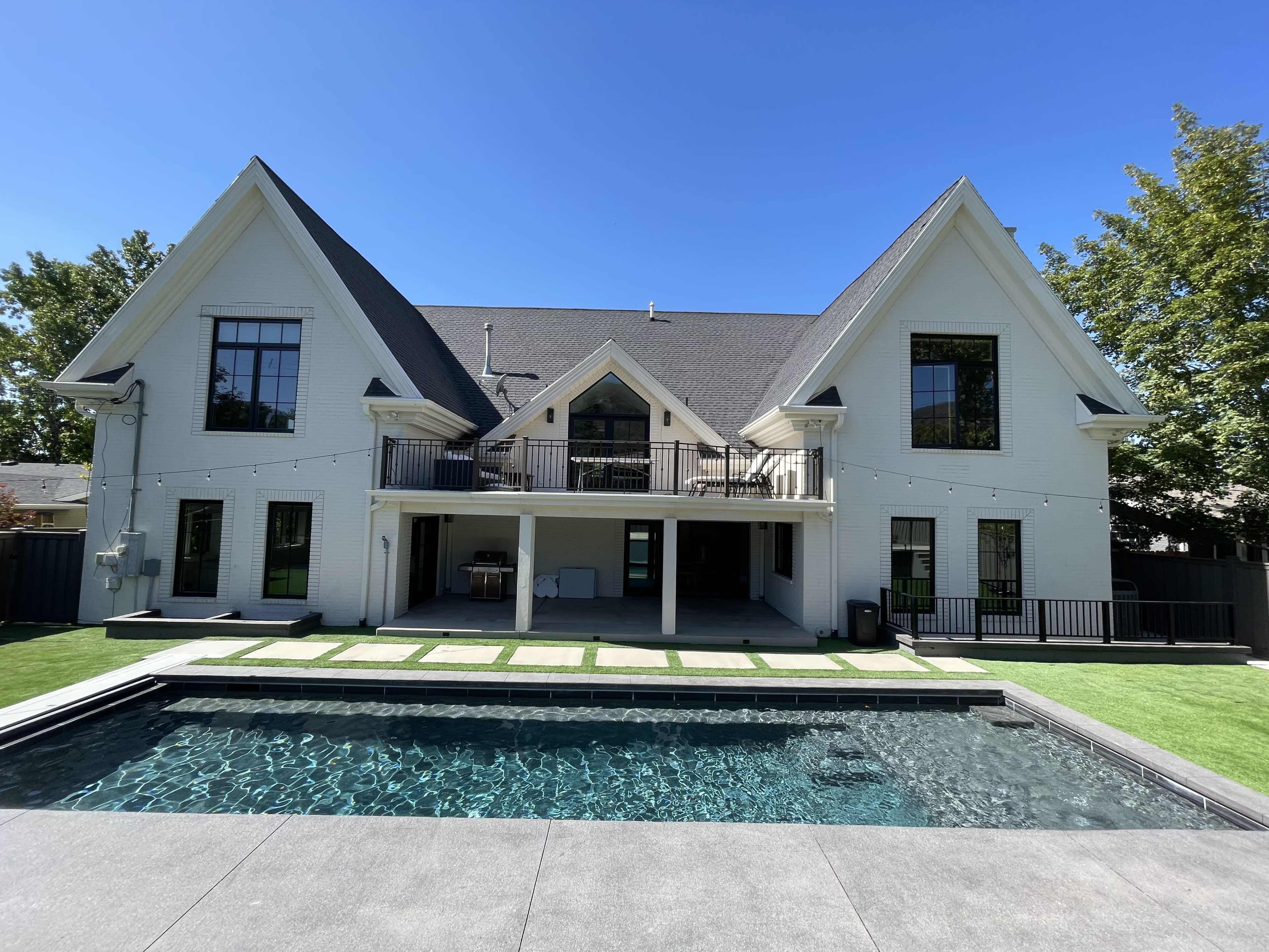 A modern two-story house with a sloped roof and large windows overlooks a rectangular pool surrounded by a stone patio and grass lawn.