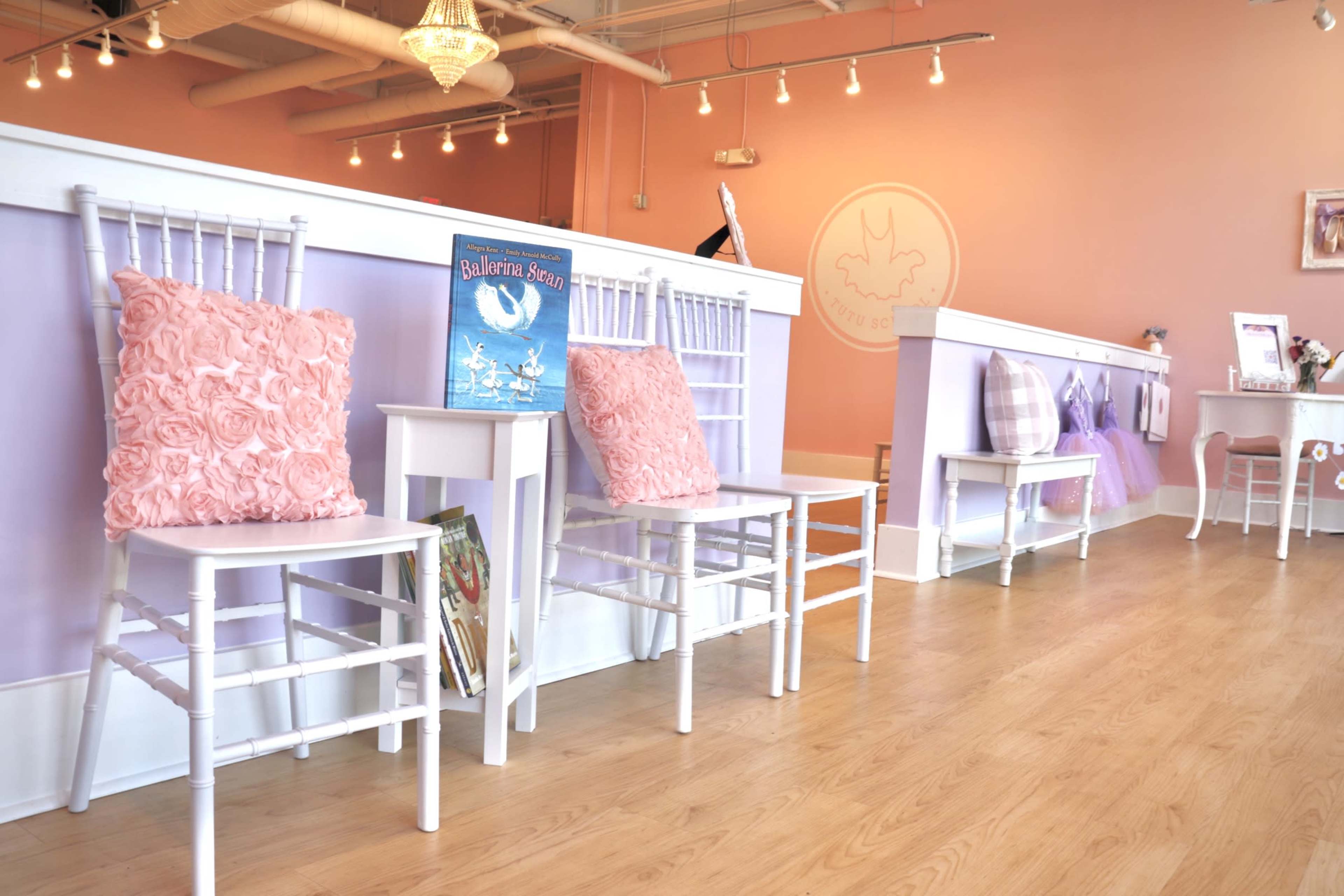 A row of white chairs with pink cushions and a decorated book sit against a light purple wall in a brightly lit room.