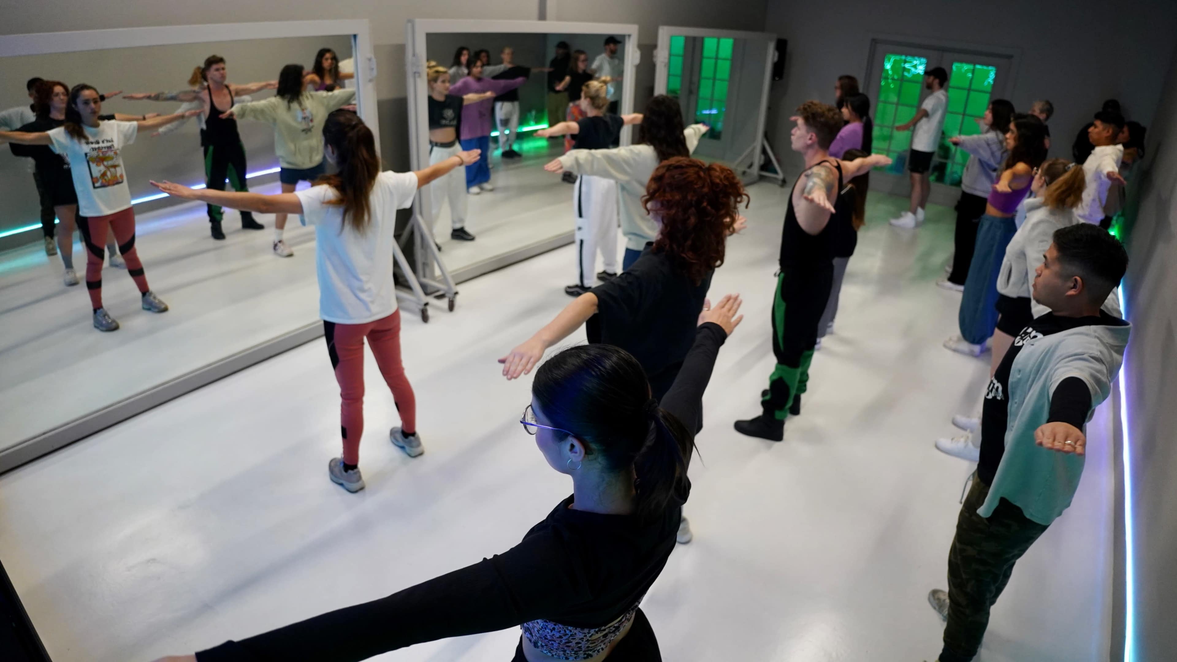 A group of people is participating in a dance class, with their reflections visible in large mirrors on the walls.