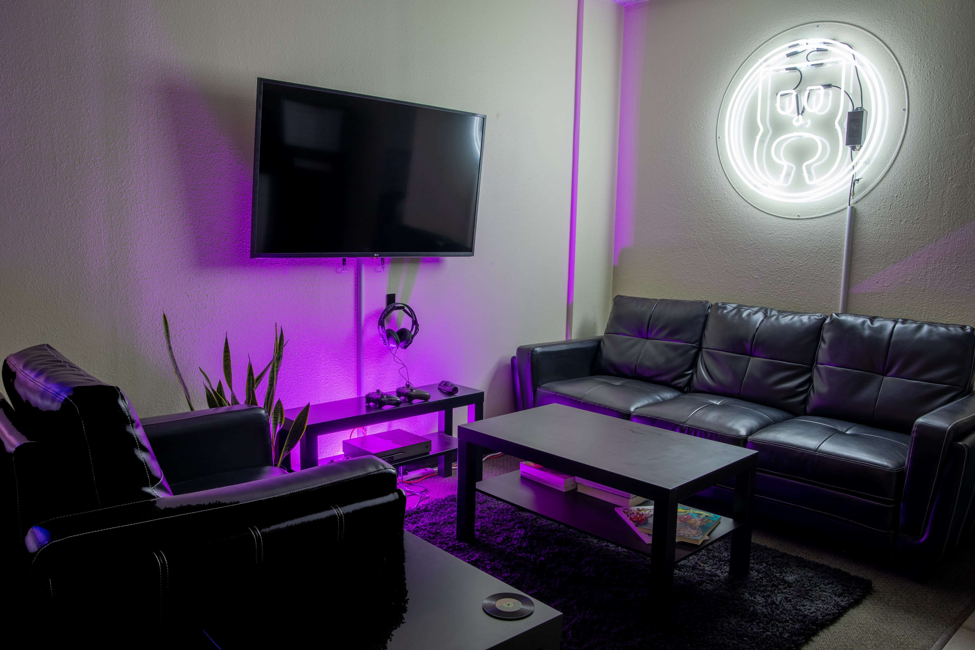 A modern living room with two black leather sofas, a large wall-mounted television, a coffee table, and a neon light fixture on the wall.