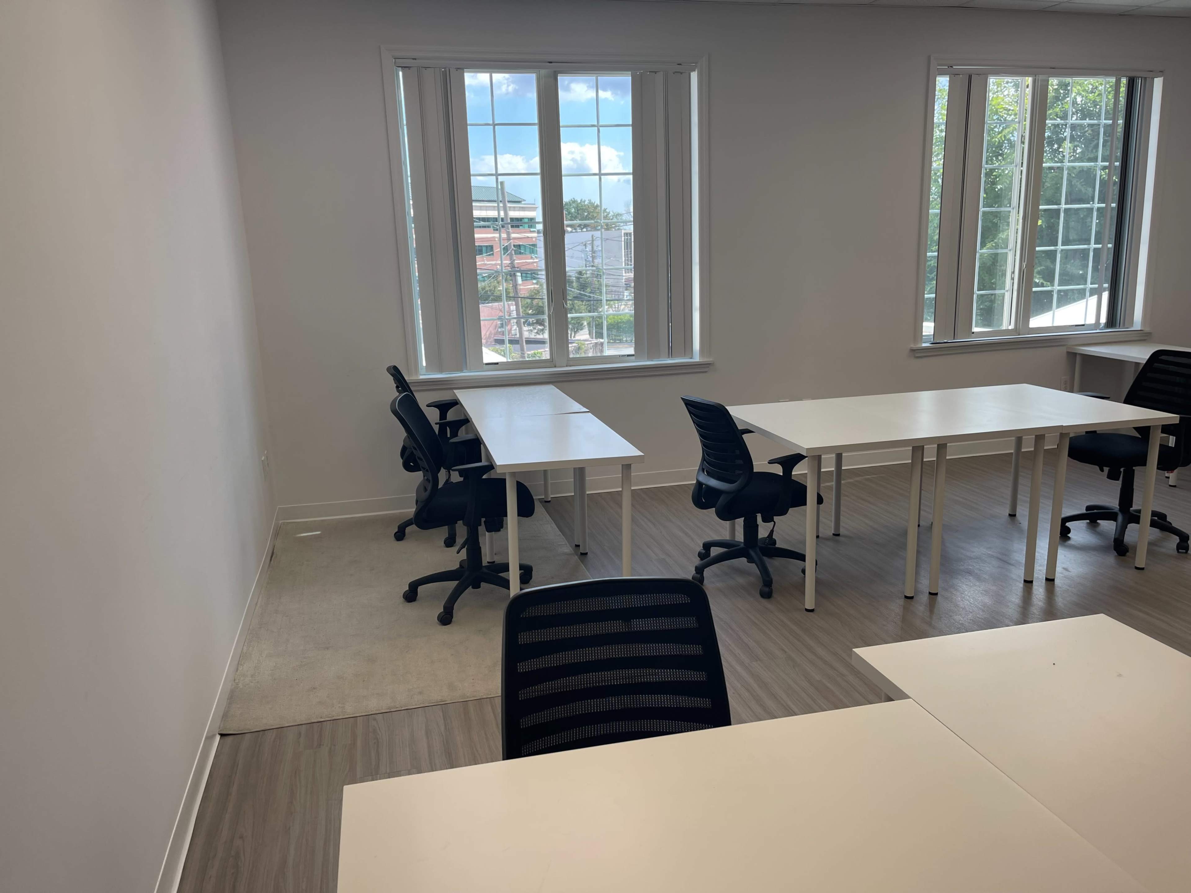 The image shows an empty office space with several desks and chairs arranged near large windows.
