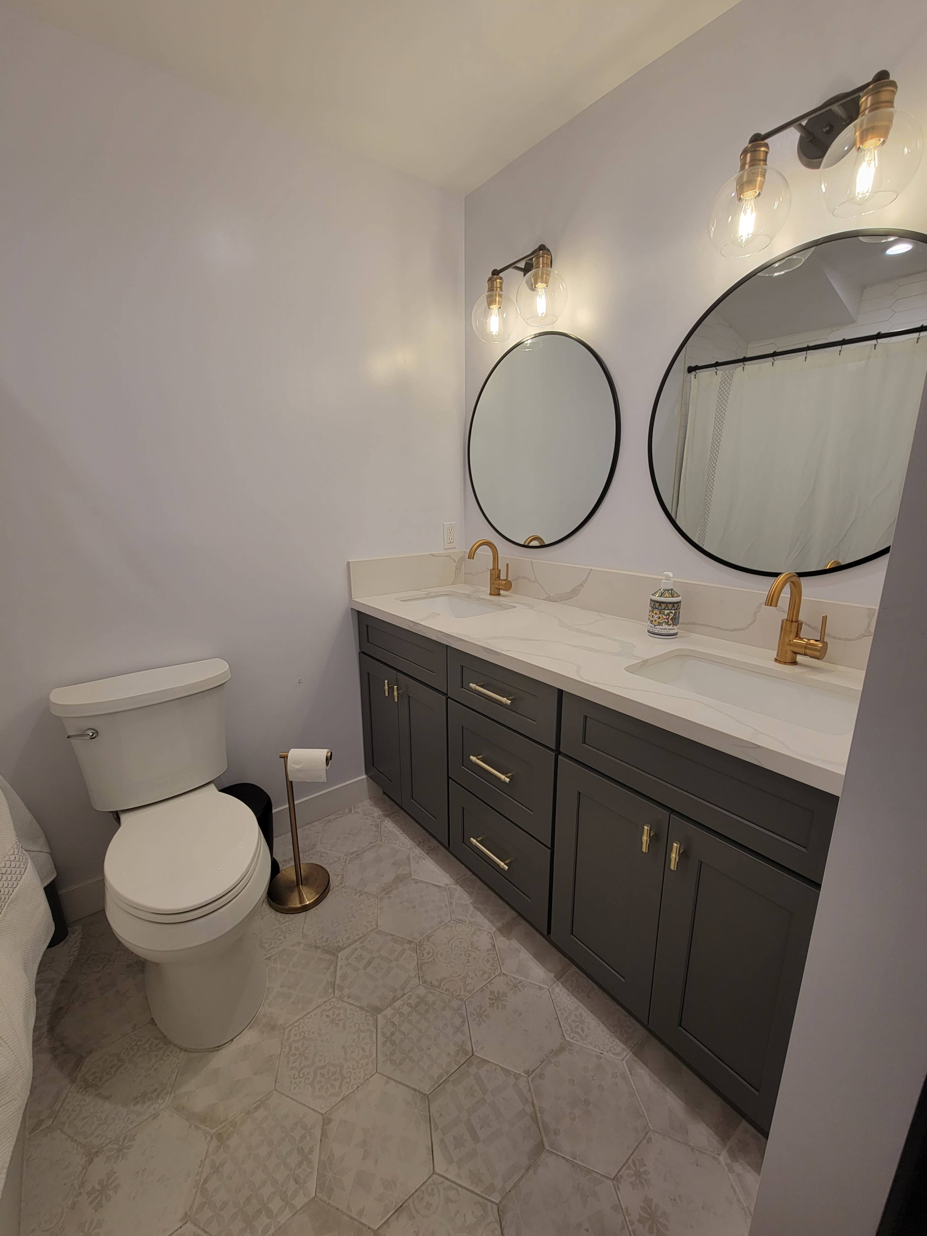 A modern bathroom with a white marble countertop, double mirrors, a dark gray vanity with gold fixtures, and a standard toilet.