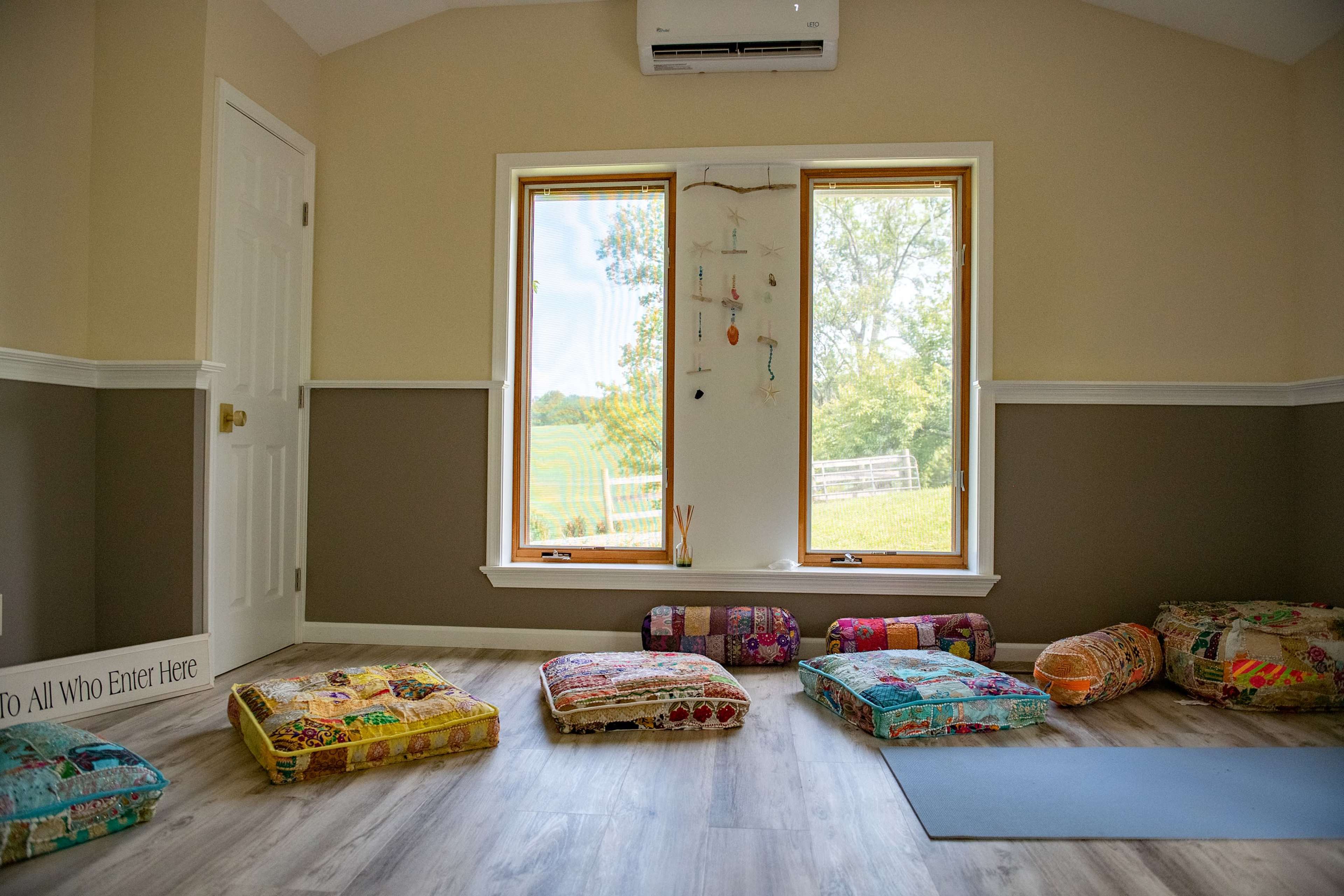 The room features colorful floor cushions arranged on a wooden floor, with two large windows offering a view of greenery outside.