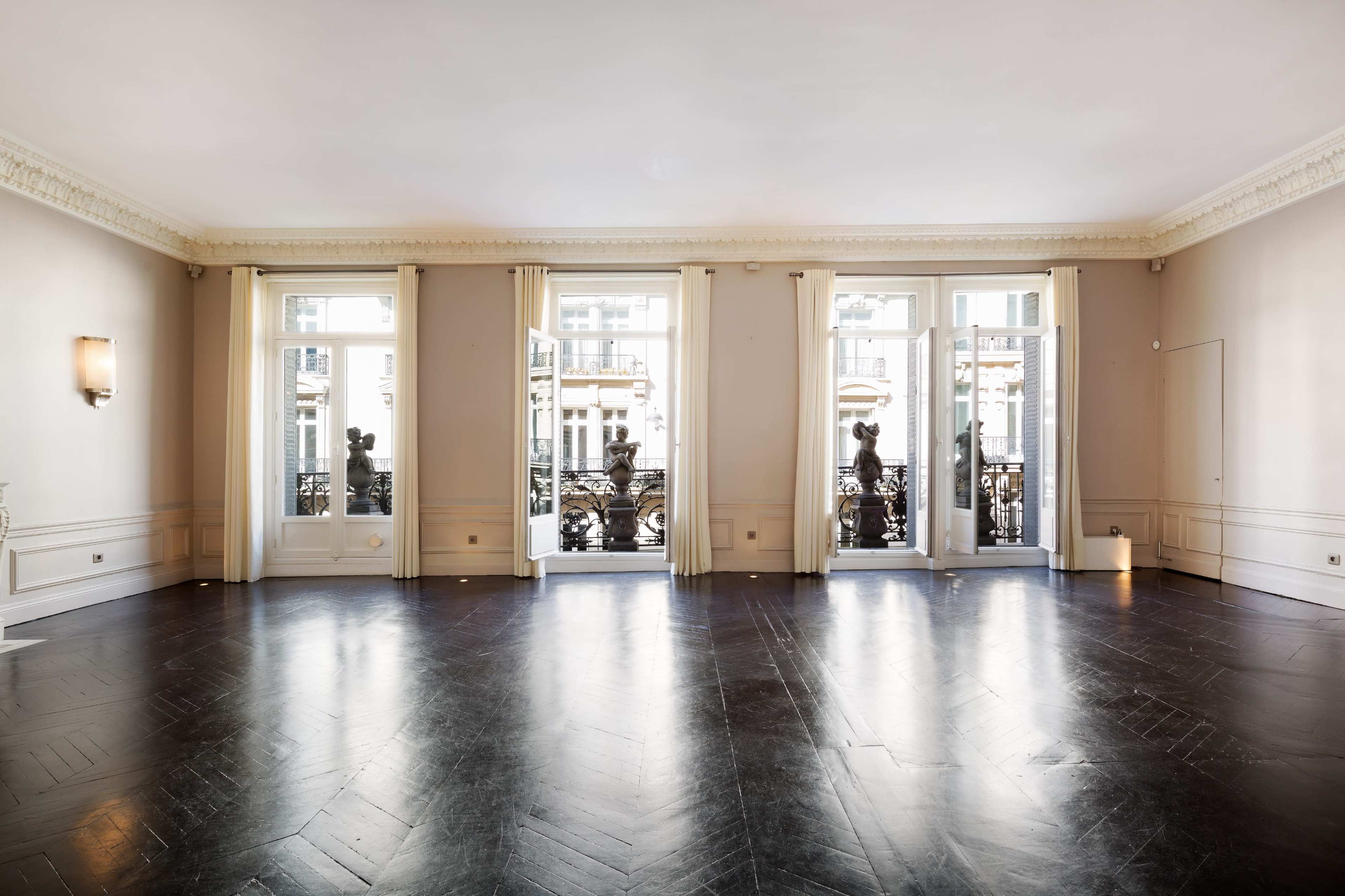 The image shows an empty room with dark wooden flooring and large windows that open to a balcony.
