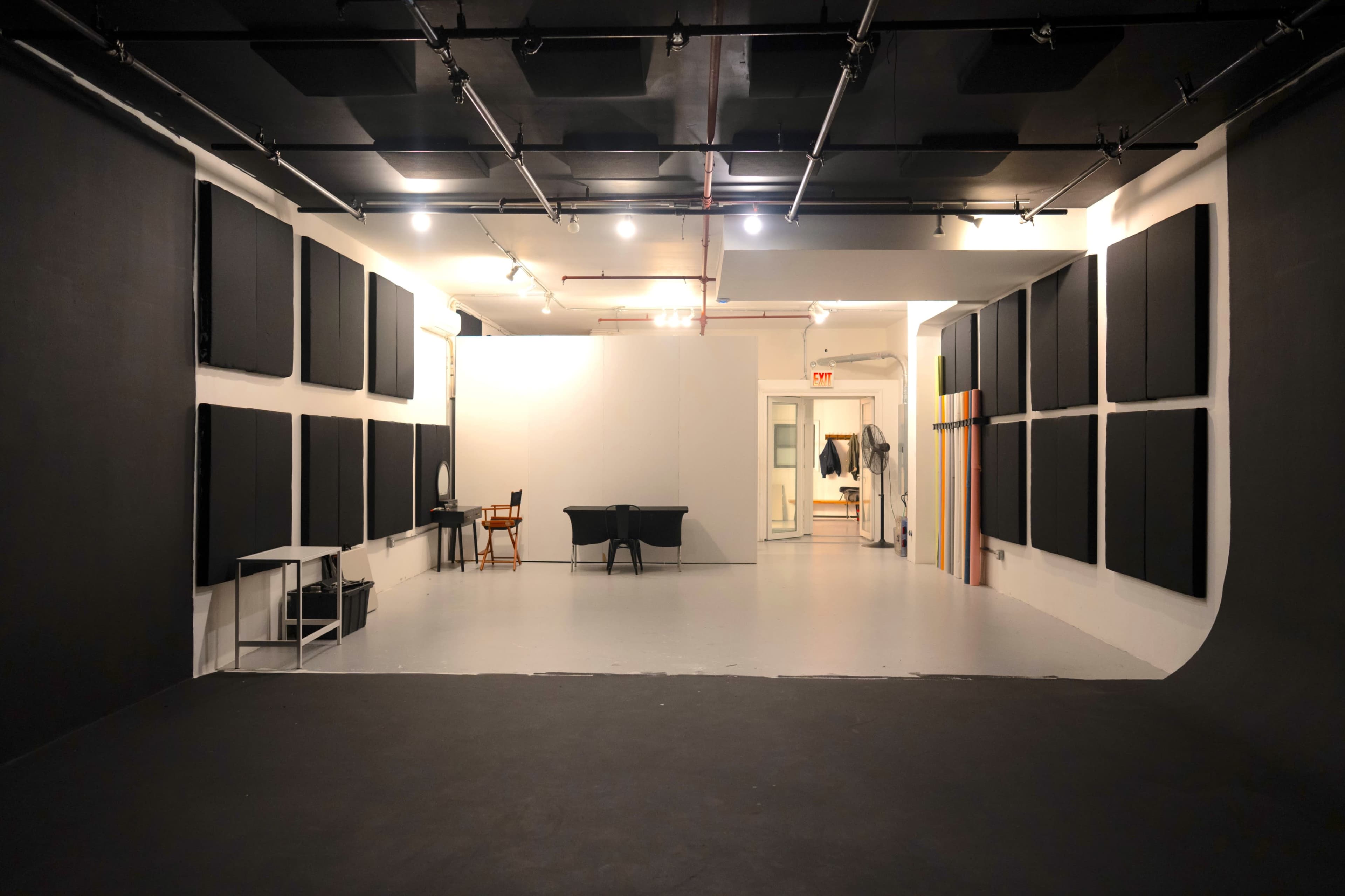 The image shows a spacious studio with black soundproofing panels on the walls, a large blank backdrop, and various pieces of furniture, including tables and chairs, in a well-lit environment.