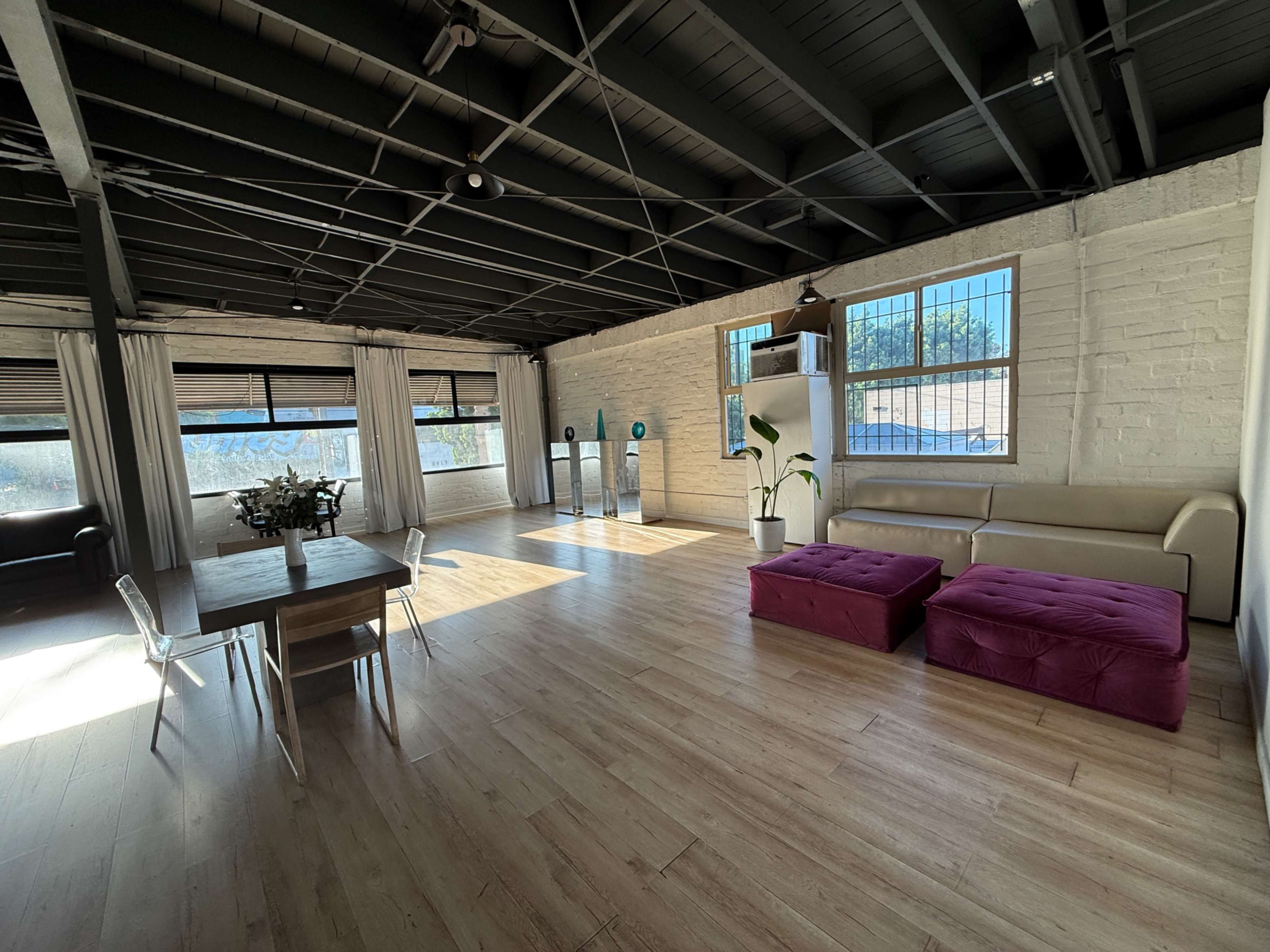 The image shows a spacious, modern interior featuring a light-colored wooden floor, a dining table with chairs, a sectional sofa, and large windows allowing natural light to enter.