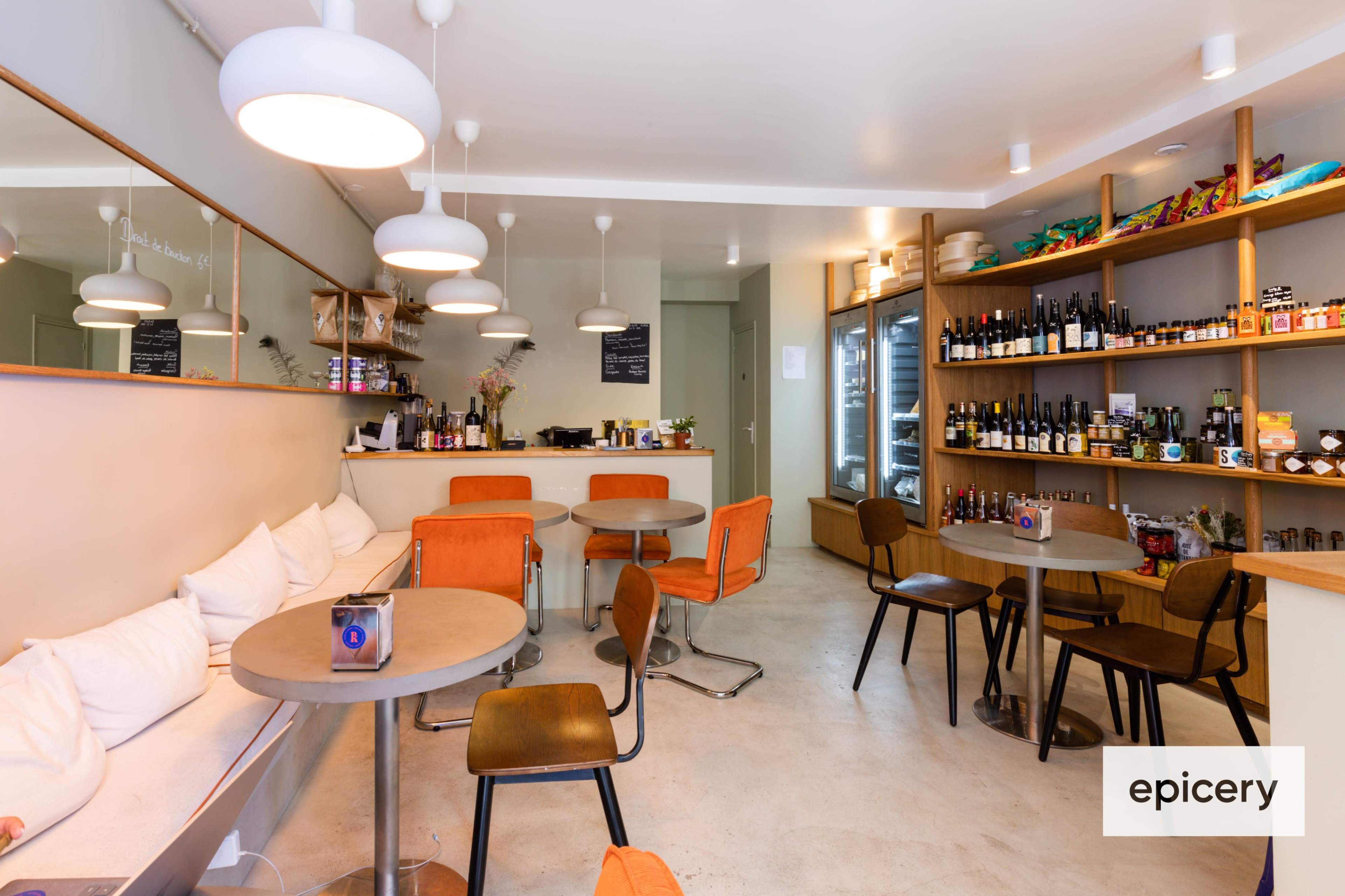 The image shows a modern cafe interior with orange chairs, wooden shelves stocked with bottles, and a mirrored wall.