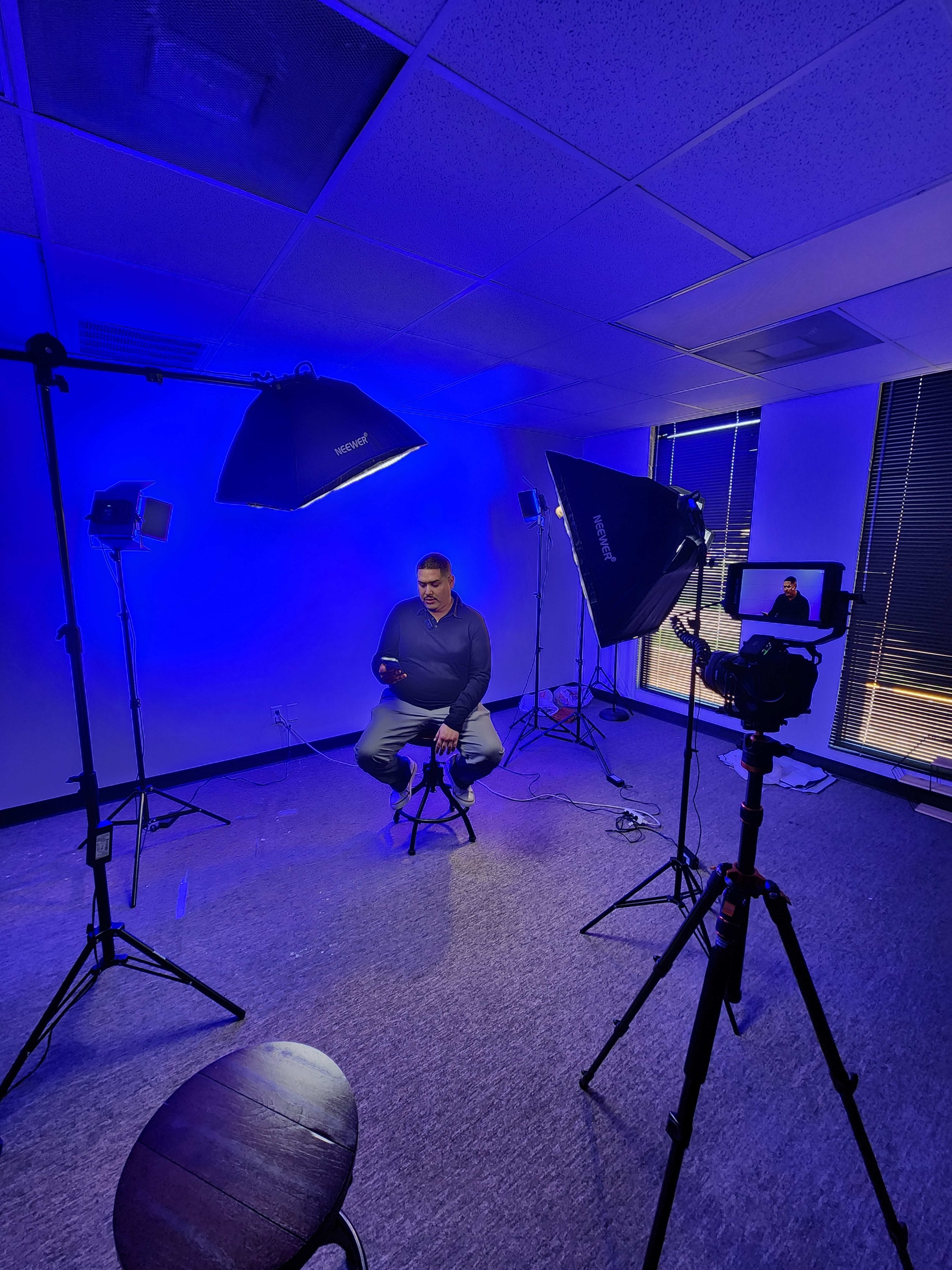 A man sits on a stool in a studio setup with various lighting equipment and a camera focused on him.
