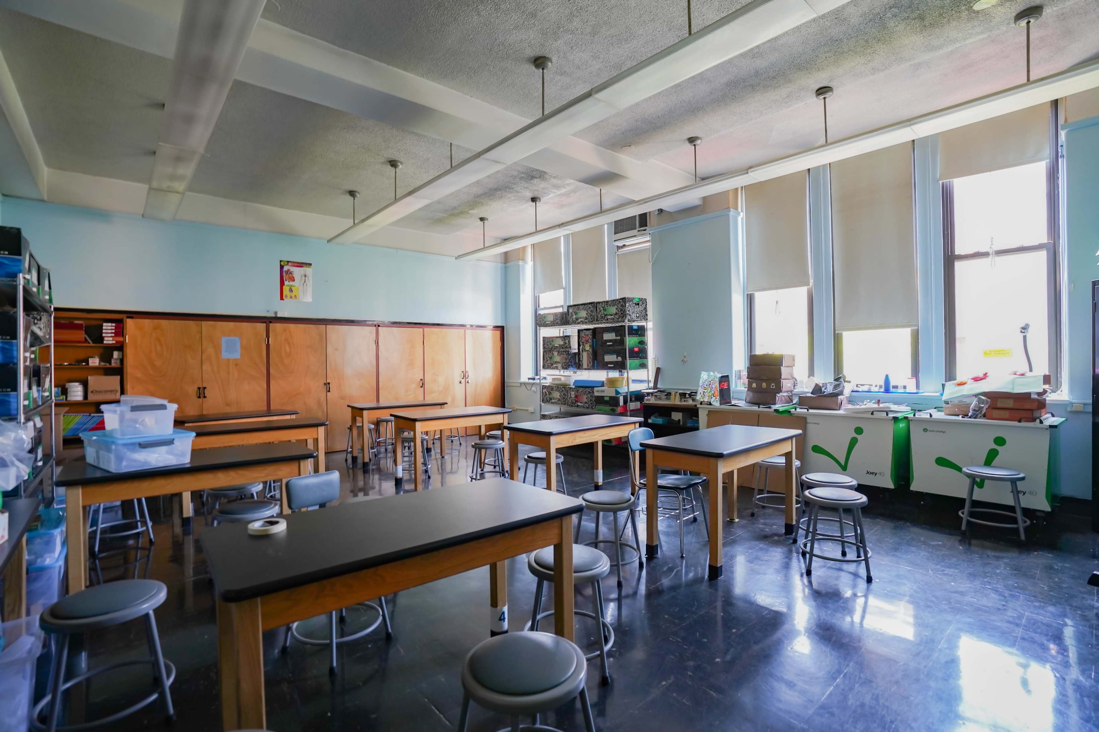 A spacious classroom with wooden tables, metal stools, and large windows, featuring storage cabinets along the walls and various supplies on counters.