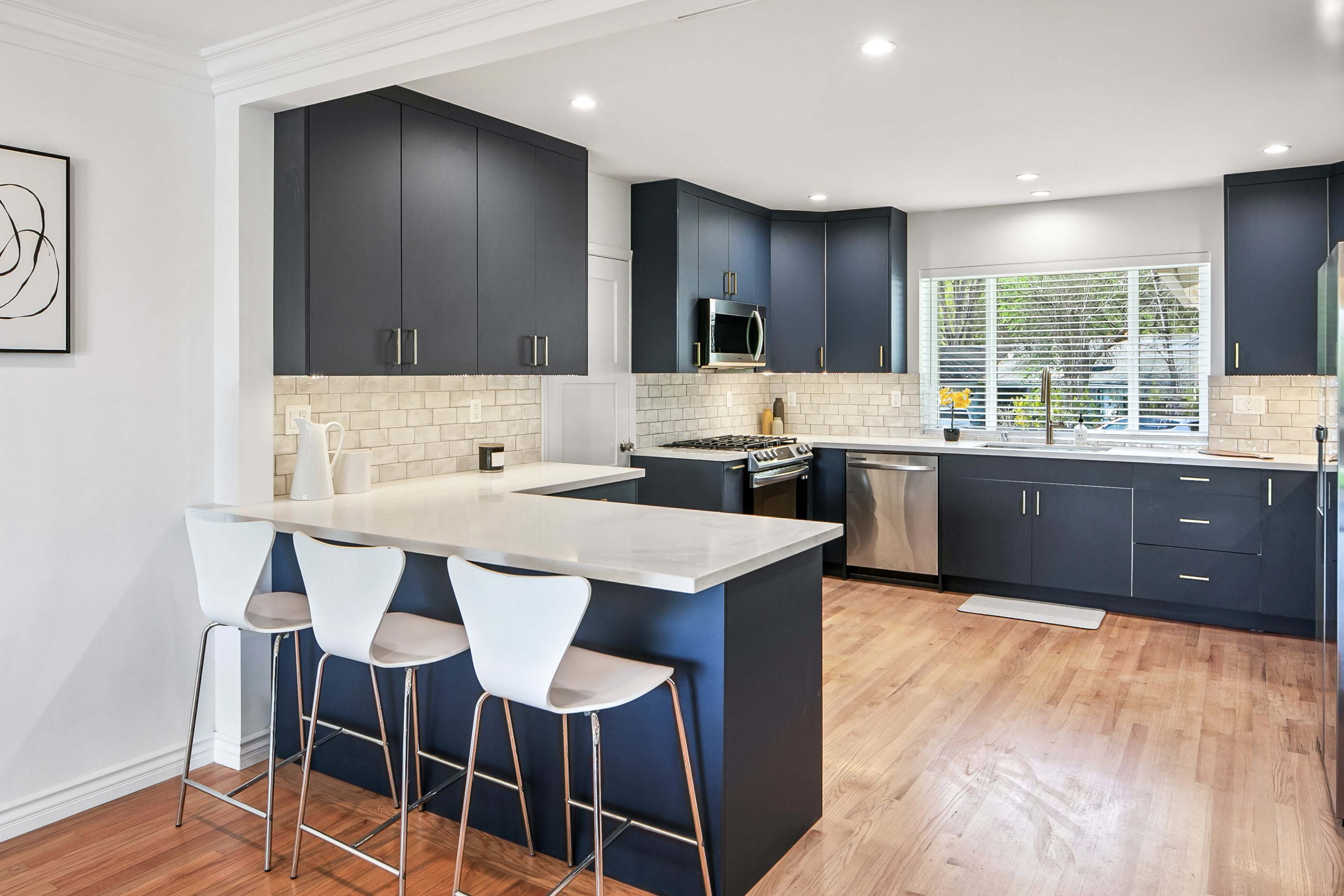 The image shows a modern kitchen with dark cabinetry, a white countertop, and stainless steel appliances, featuring a breakfast bar with three white chairs.