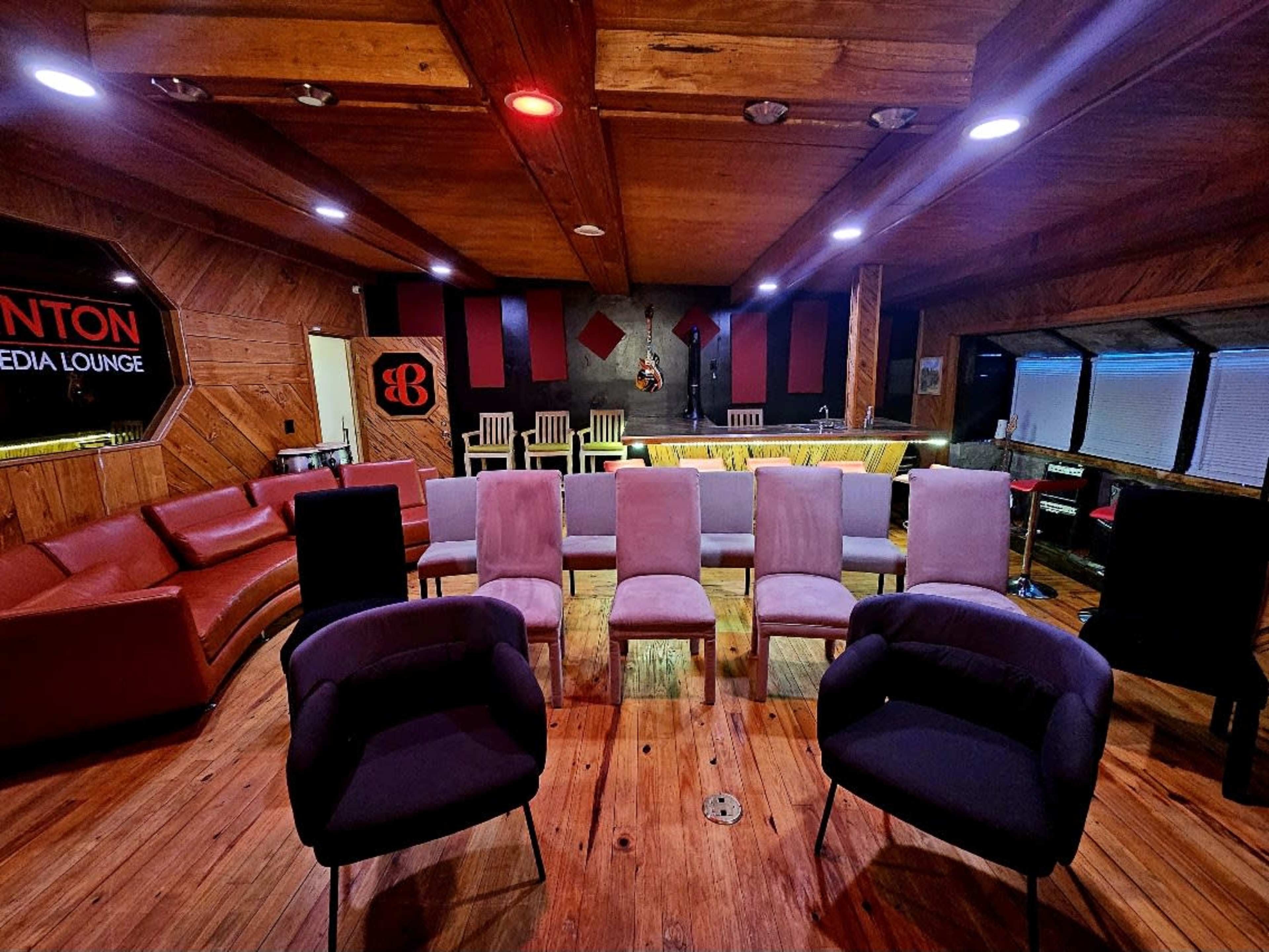 The image shows a media lounge with a wooden interior, featuring a semicircular arrangement of chairs, a bar area, and colorful lighting.