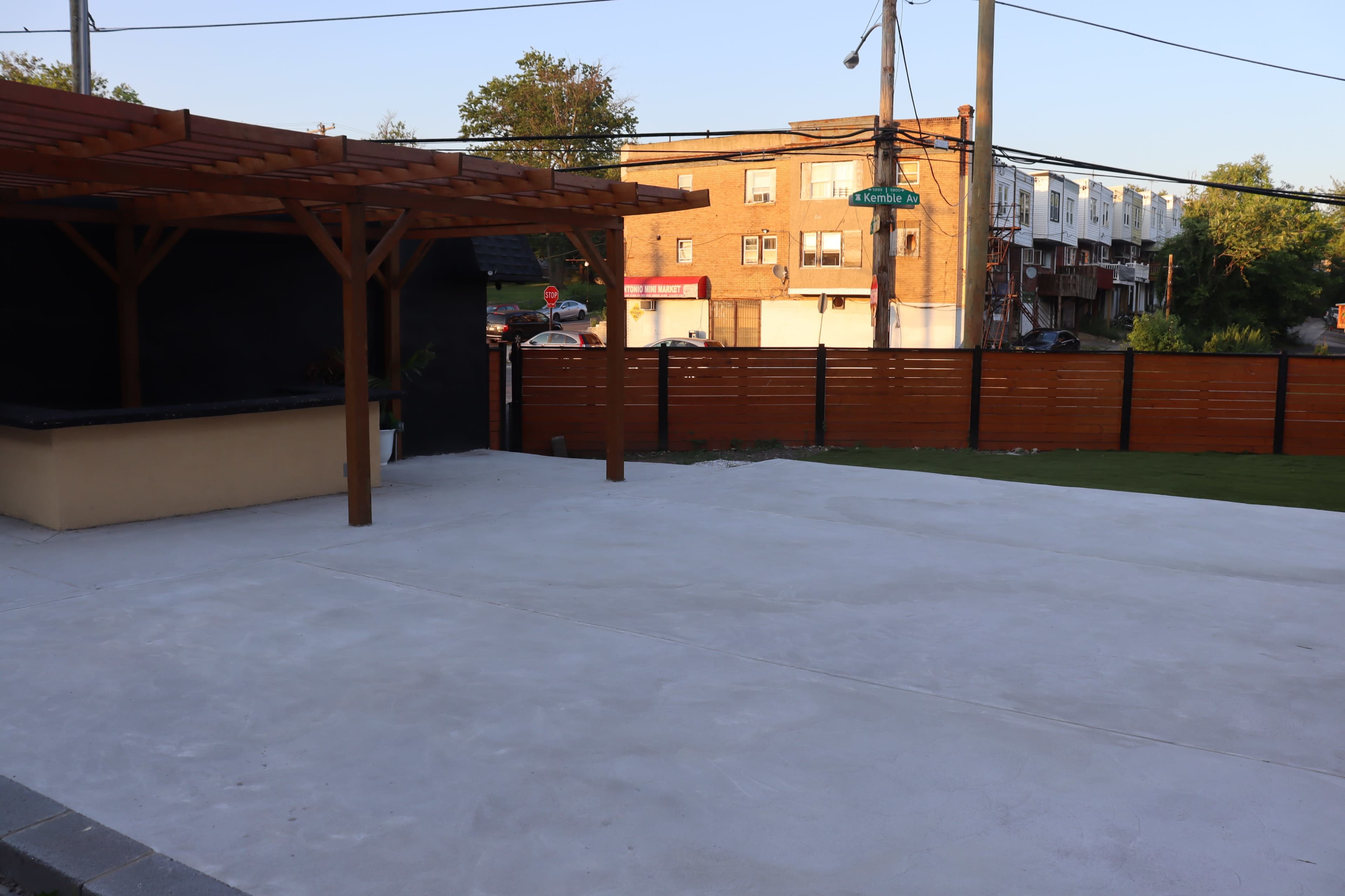 The image shows a partially finished outdoor patio area with a wooden pergola and a fence, overlooking a street with parked cars and buildings in the background.