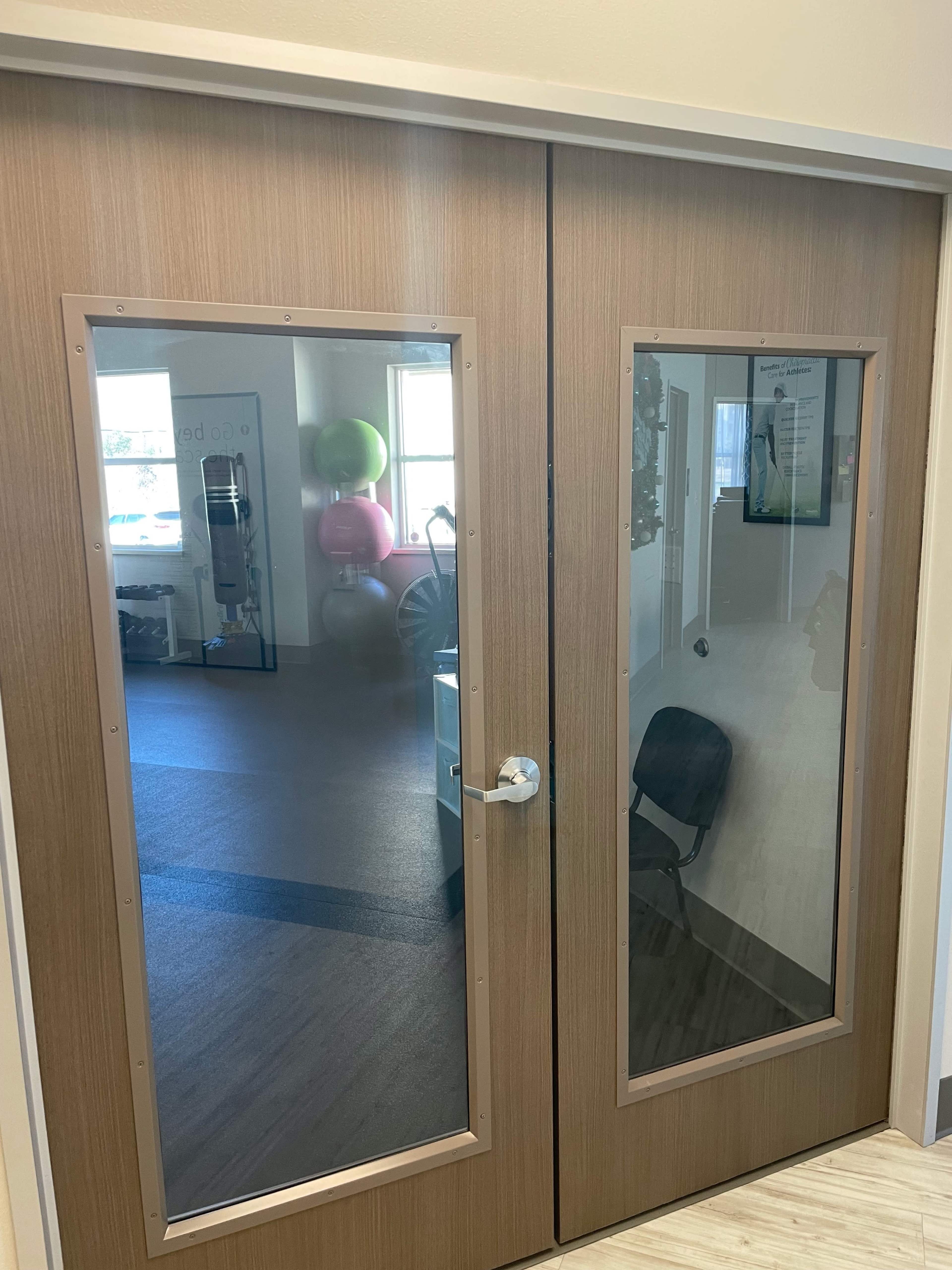 A pair of wooden double doors with glass panels, leading into a brightly lit room containing exercise equipment and fitness balls.