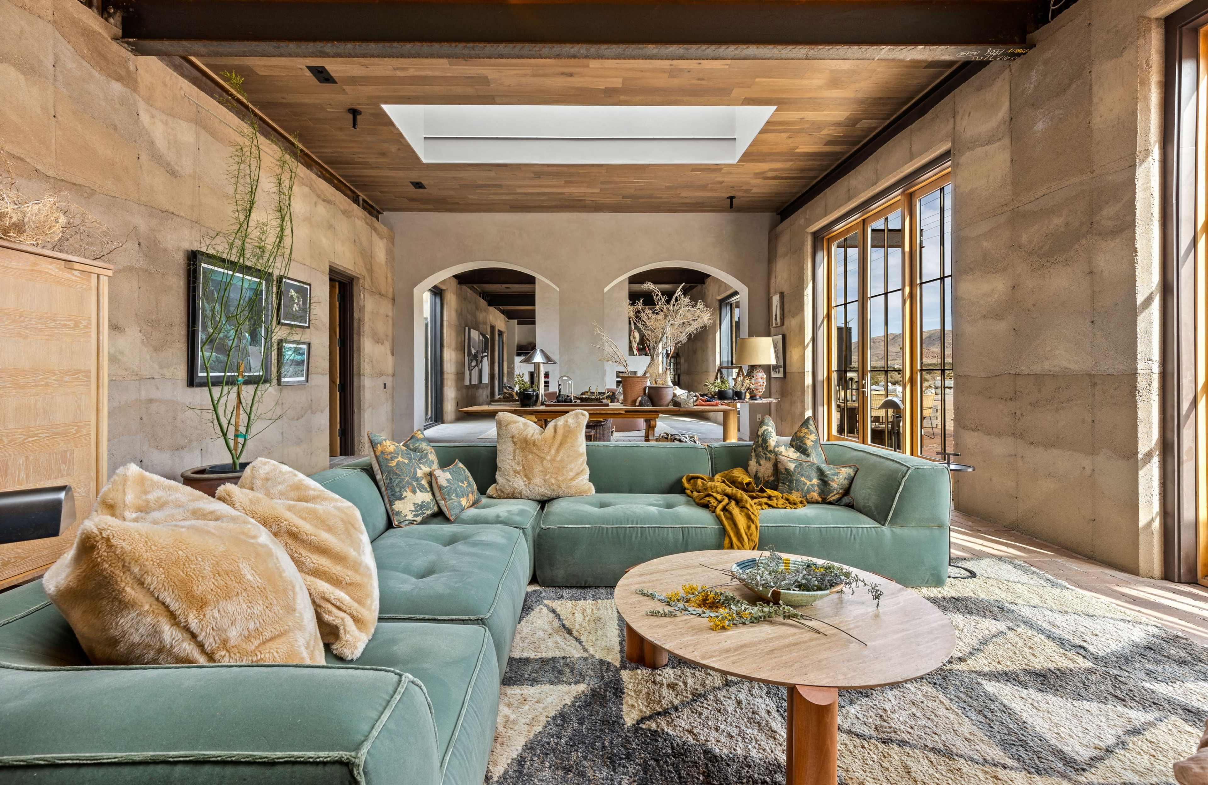 The image shows a spacious living room featuring a green sectional sofa, a wooden coffee table, and large windows that provide natural light.