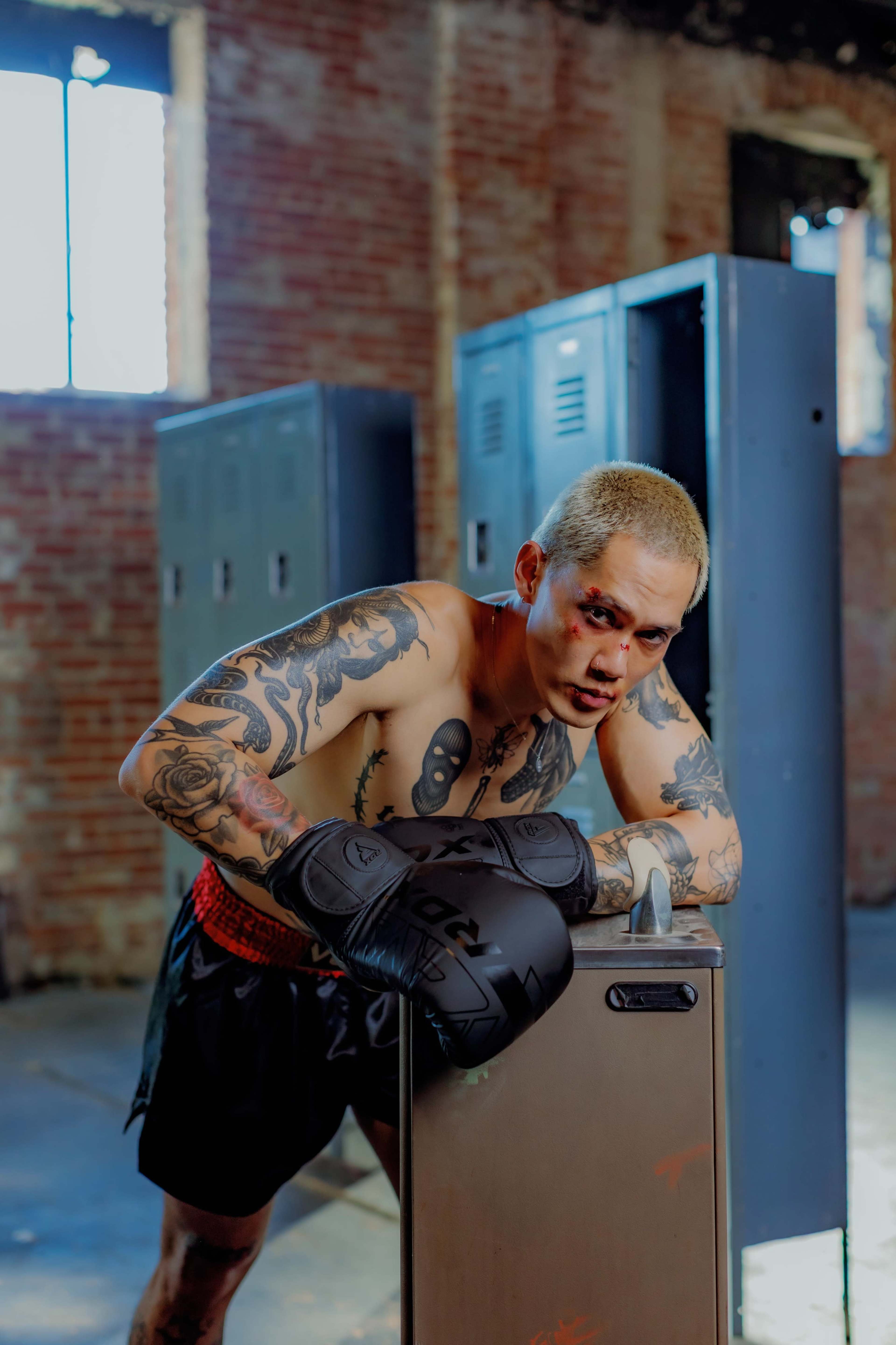 A shirtless boxer with tattoos and bloodied facial bruises leans on a locker in an industrial gym setting.