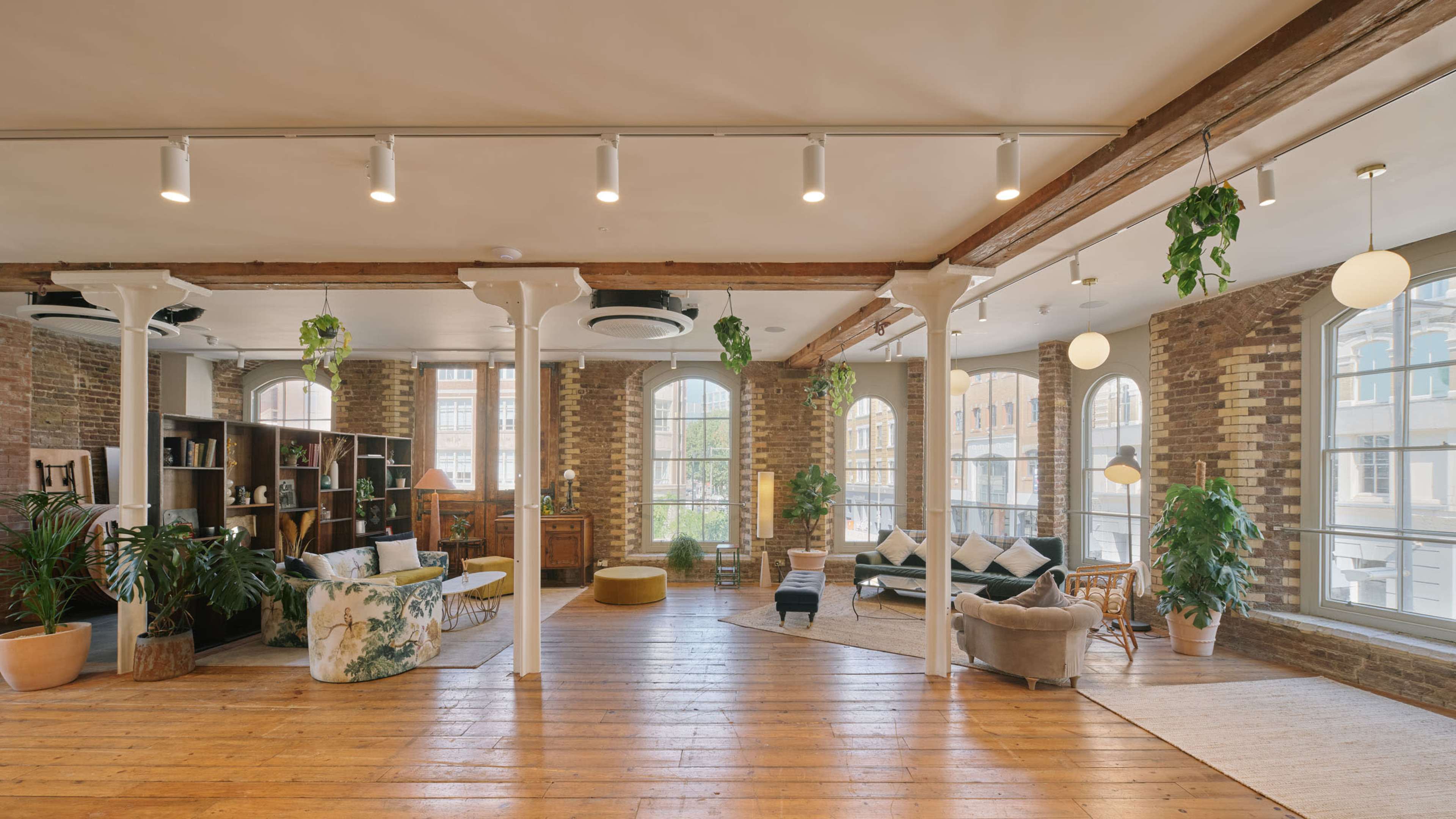 The image shows a spacious, well-lit interior featuring wooden floors, large windows, and a mix of seating areas with plants and decorative furniture.