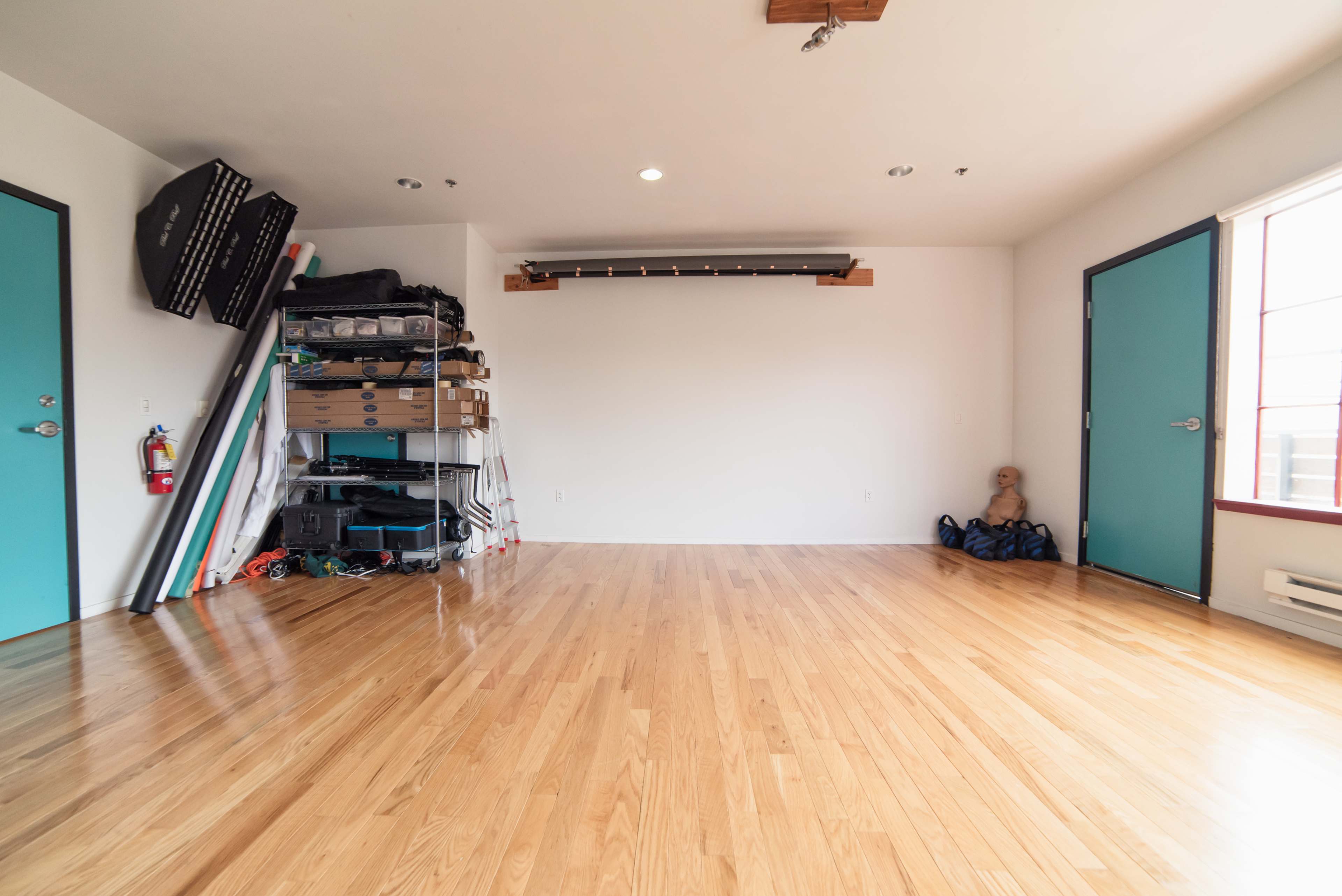 A spacious, empty room featuring light wooden flooring, turquoise doors, and storage shelves with various items along one wall.