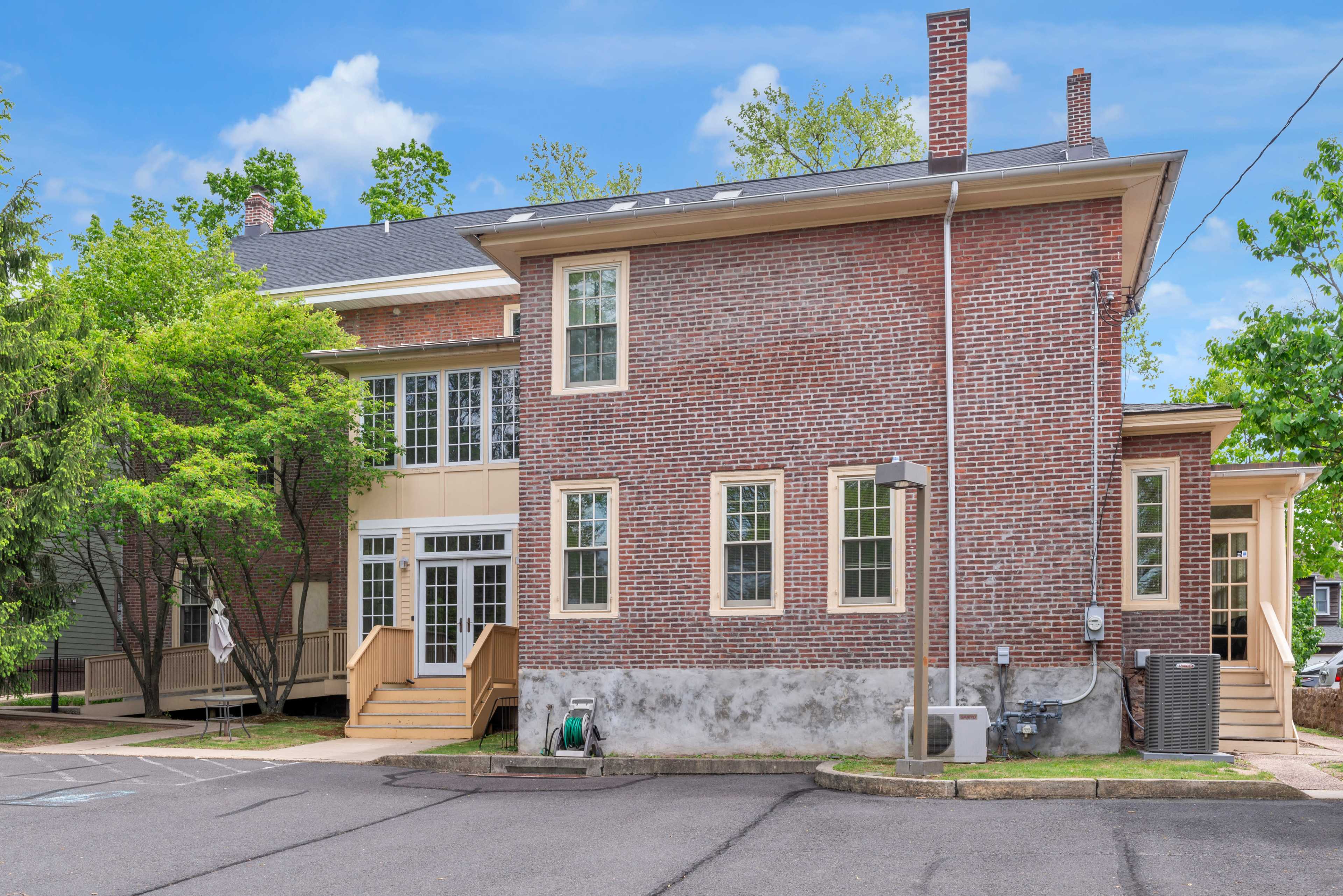 The image shows a two-story brick building with a modern addition, surrounded by trees and a paved area.