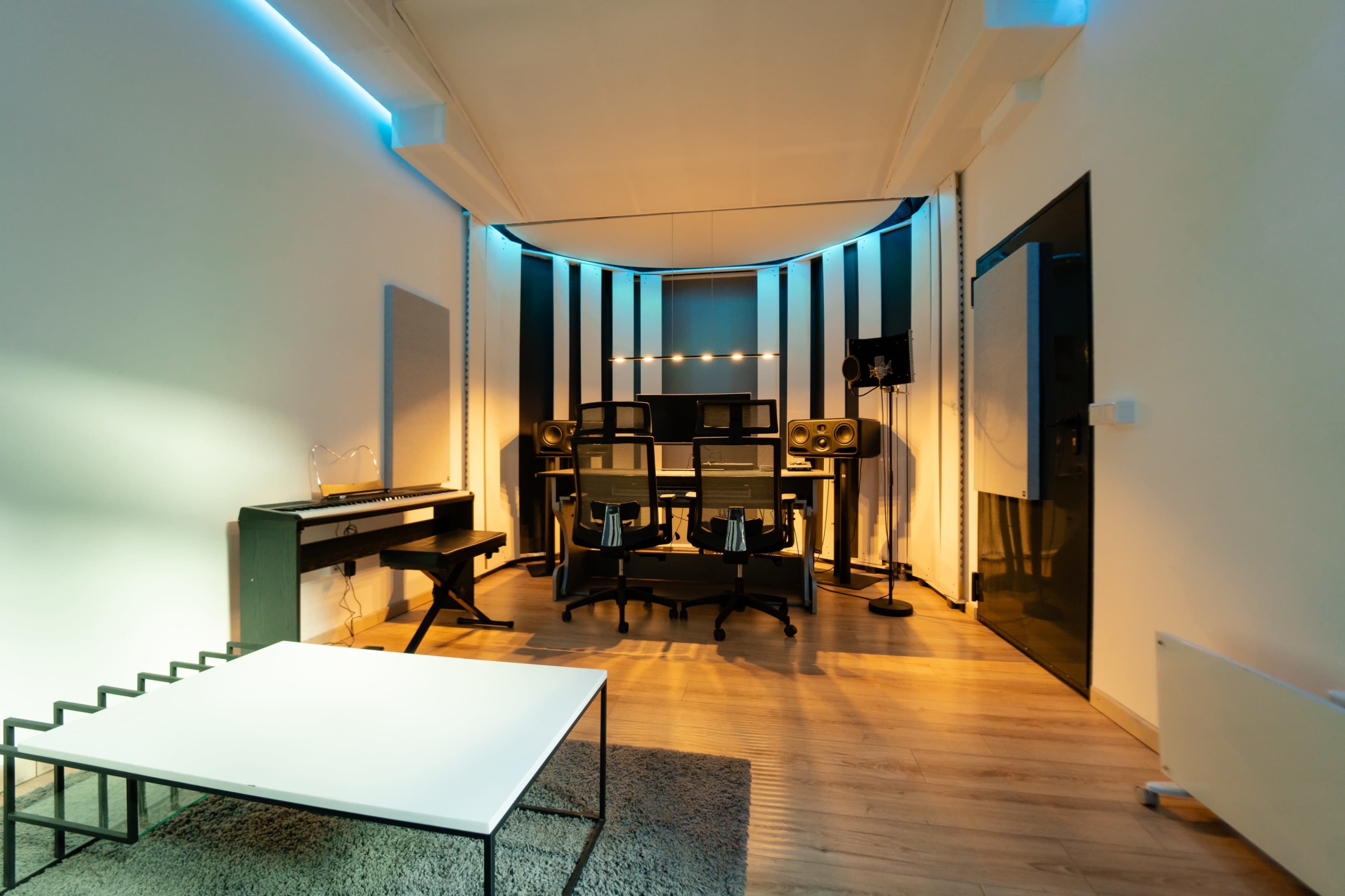 The image shows a modern recording studio featuring a desk, two chairs, sound equipment, and ambient lighting along the walls.