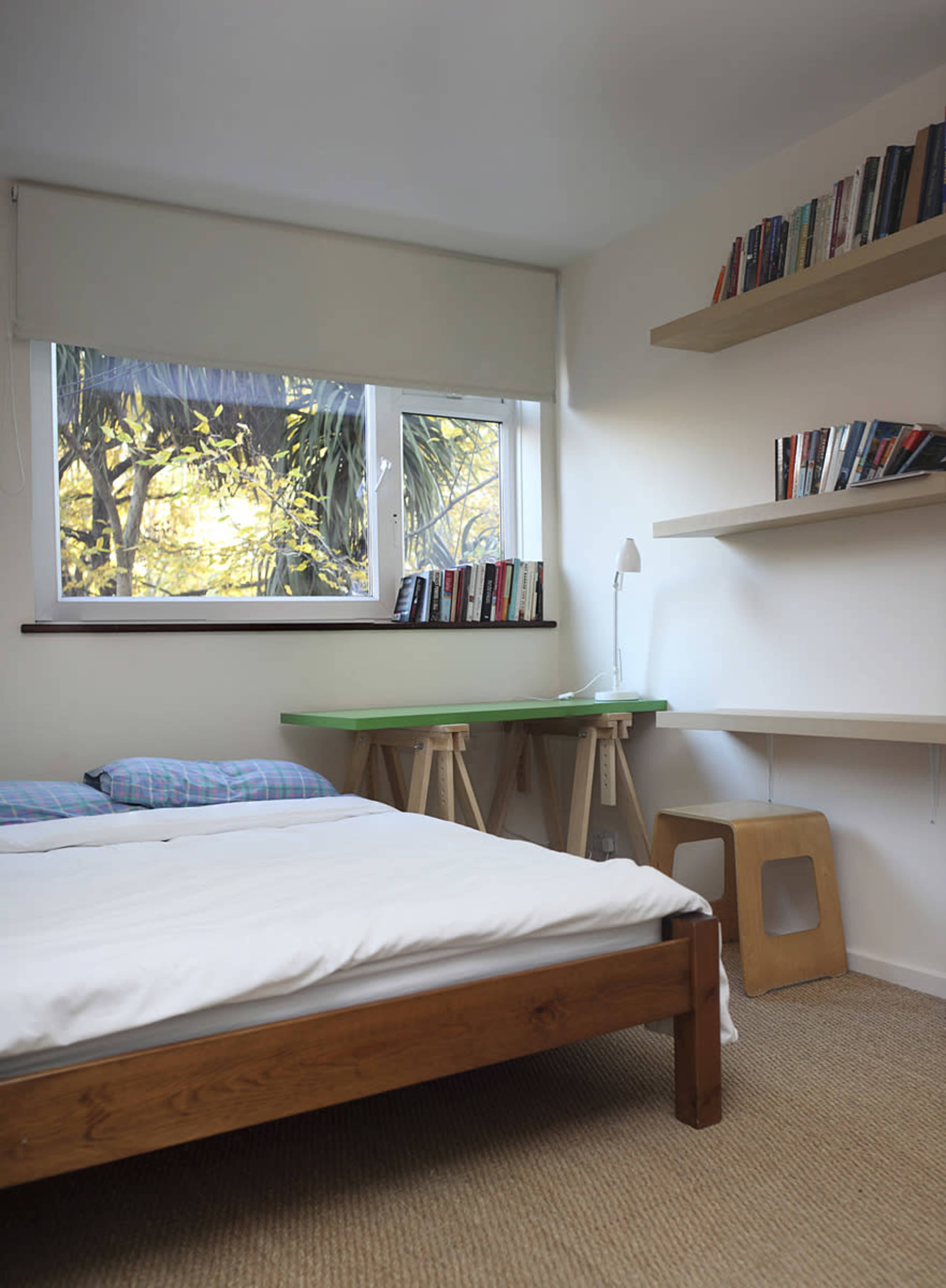 A simple bedroom with a wooden bed, a green desk, a bookshelf on the wall, and a window letting in natural light.