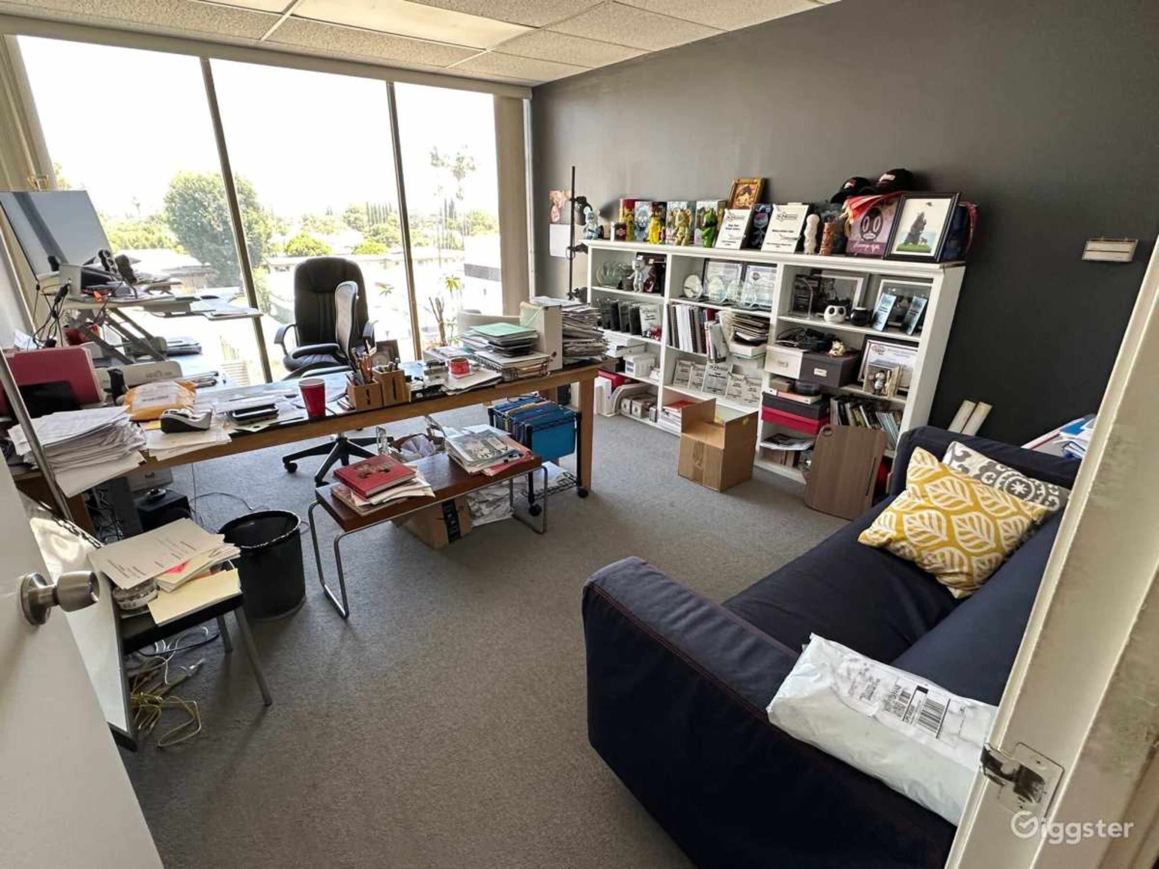 A cluttered office space with a large desk, shelves filled with boxes and books, a computer monitor, and a black chair, all illuminated by natural light from a large window.