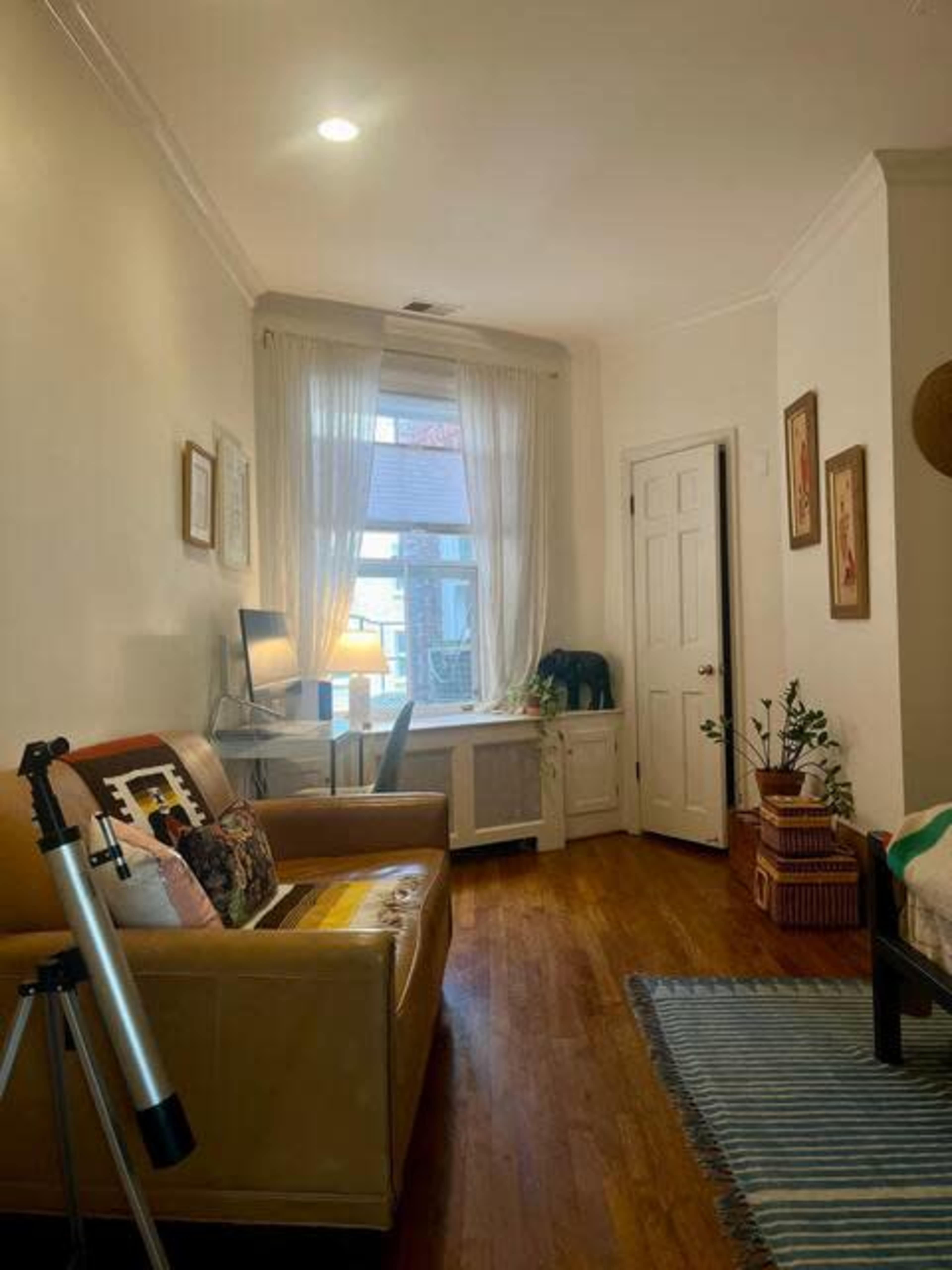 A small, well-lit room featuring a desk by a window, a couch with a blanket, and a bed with a striped rug on the floor.