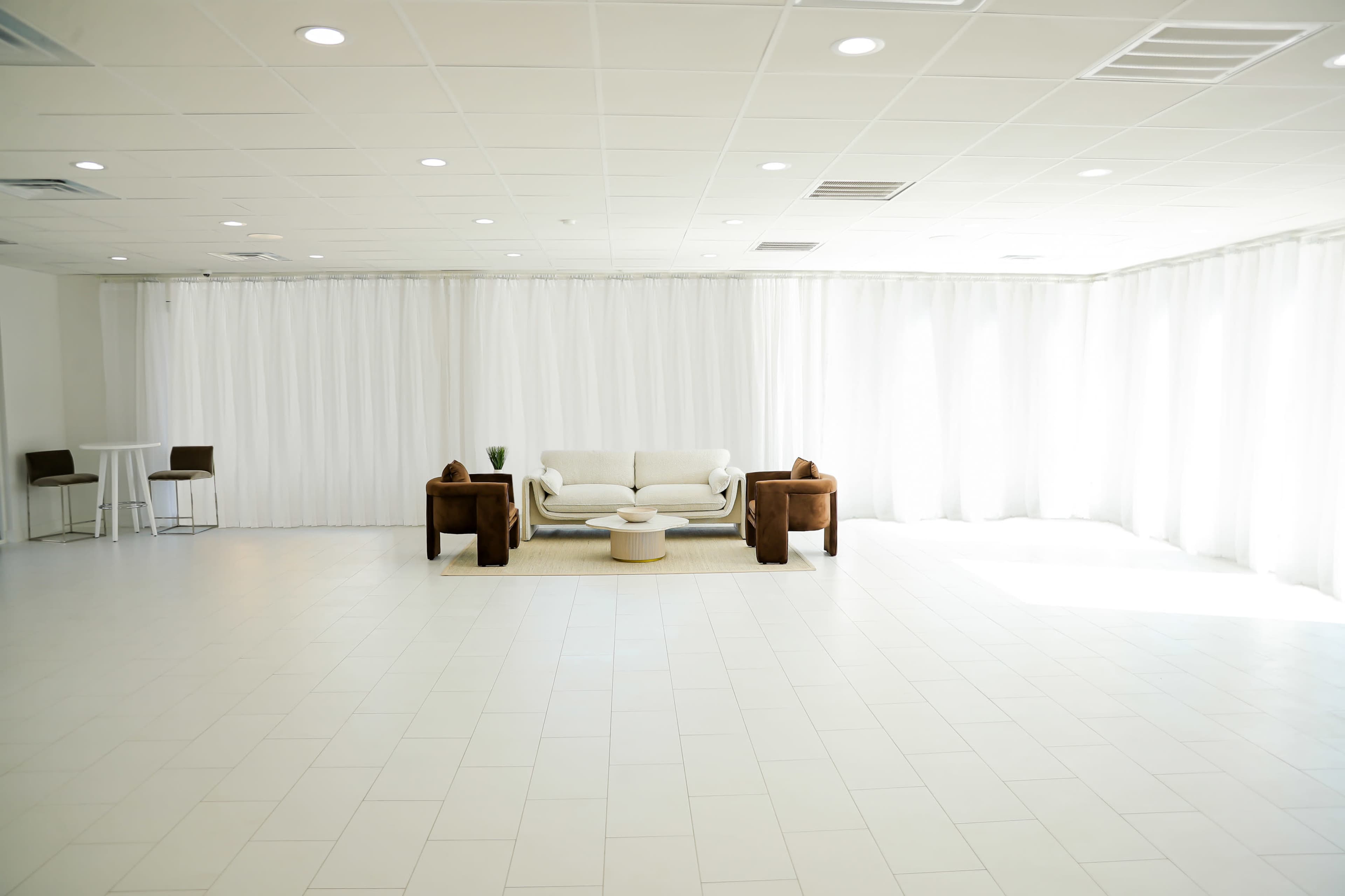 A spacious, well-lit room featuring a beige sofa, two brown armchairs, a round coffee table, and sheer white curtains lining the walls.