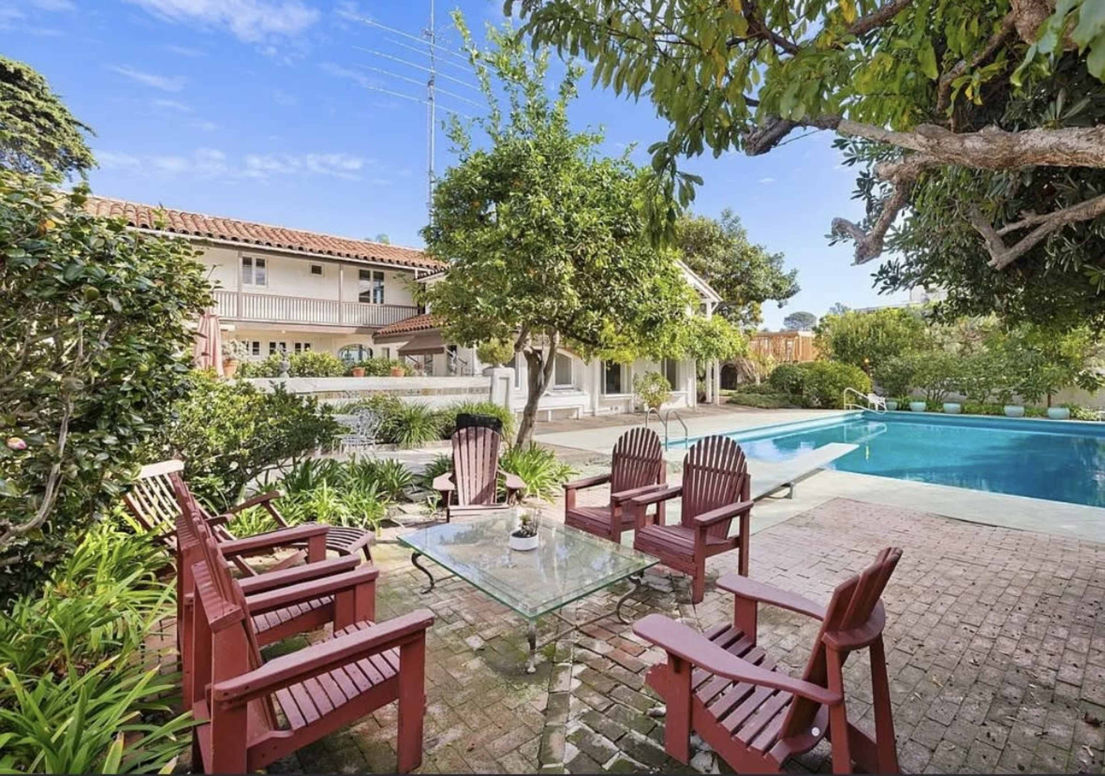 A backyard features a swimming pool surrounded by landscaped greenery and a seating area with red wooden chairs and a glass table.
