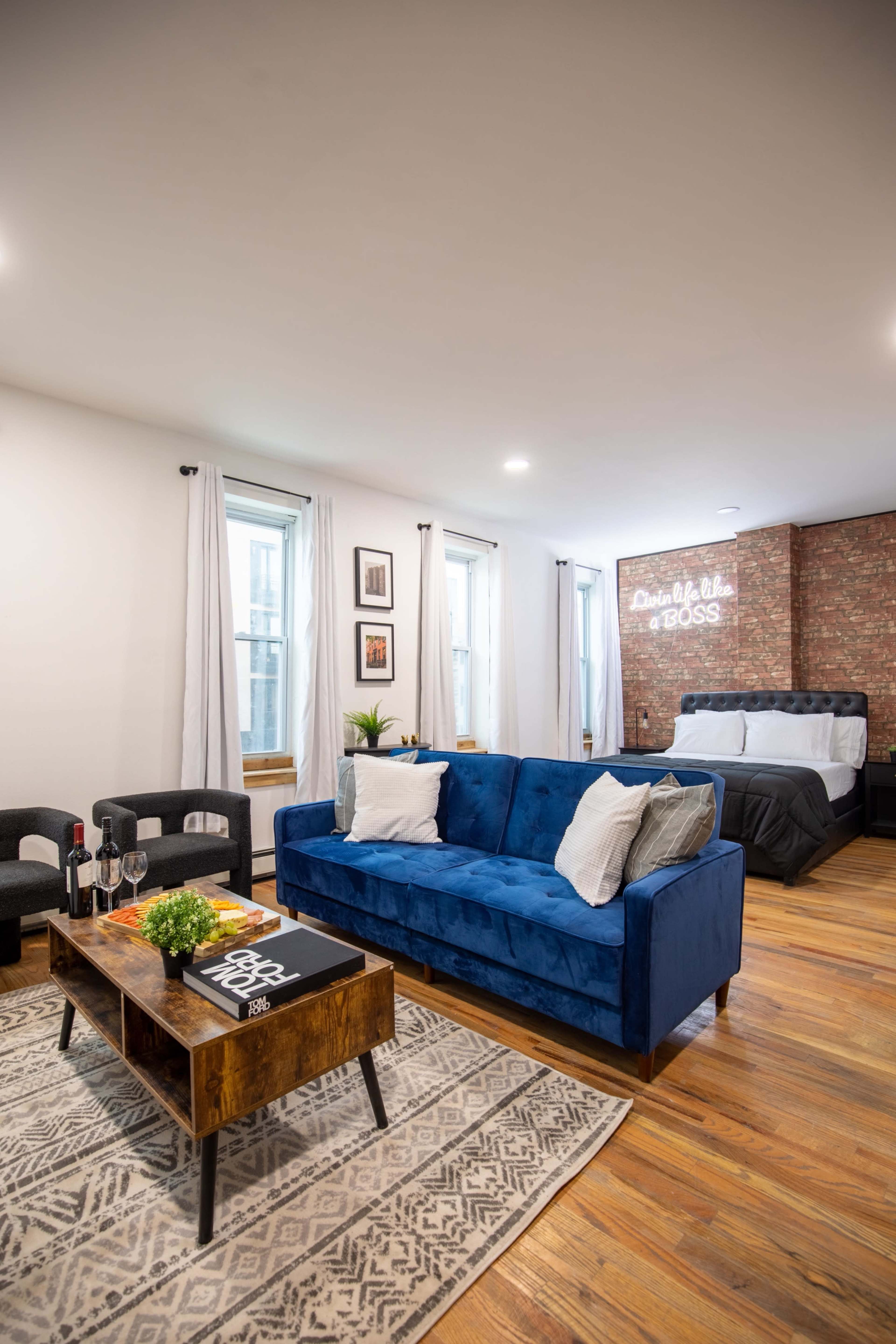 A stylish studio apartment featuring a blue couch, a wooden coffee table, and a bed against a brick wall with decorative lighting.