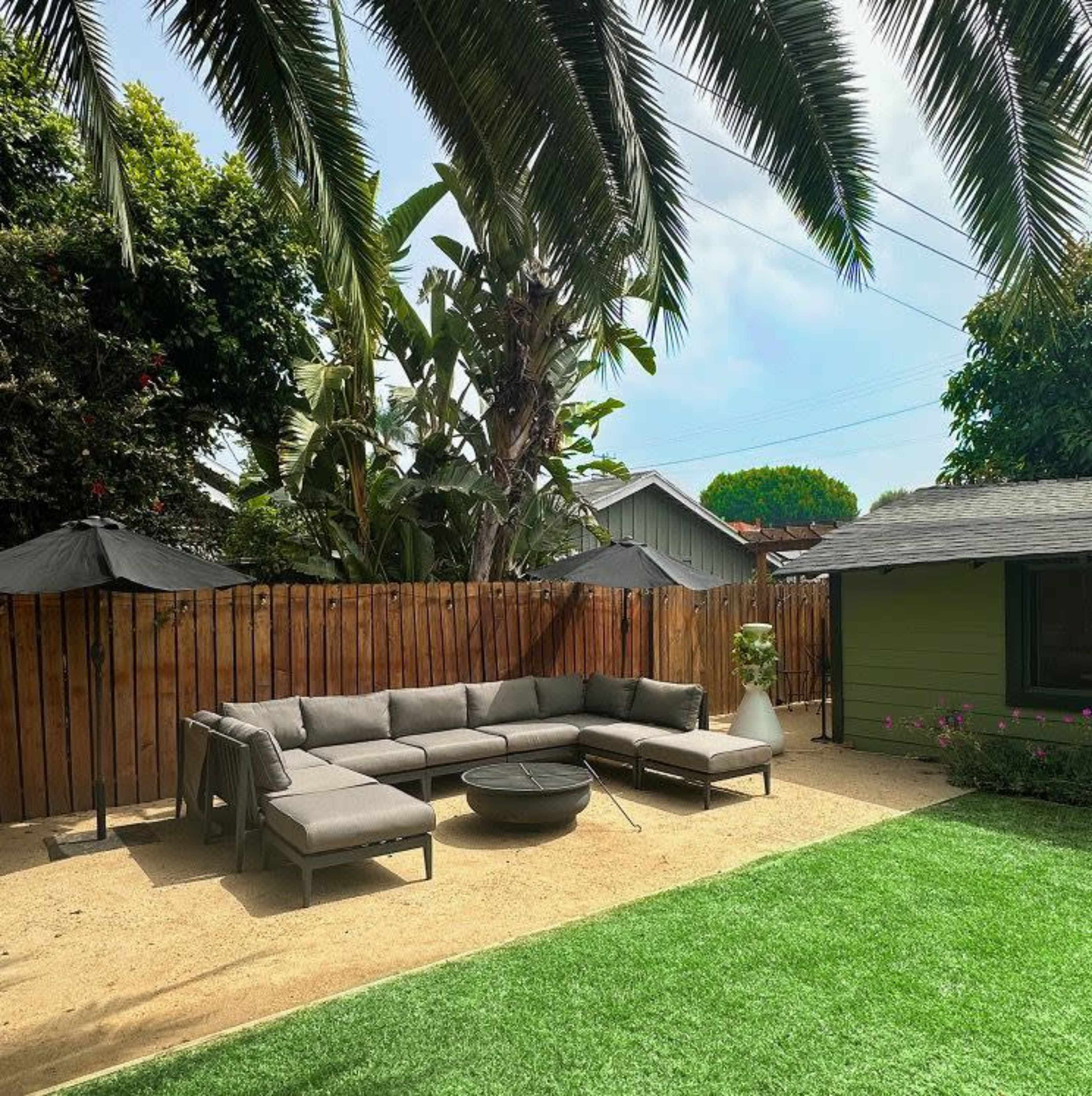 The image shows a backyard with a gray sectional sofa, a circular coffee table, two black umbrellas, and a small shed, surrounded by palm trees and a wooden fence.