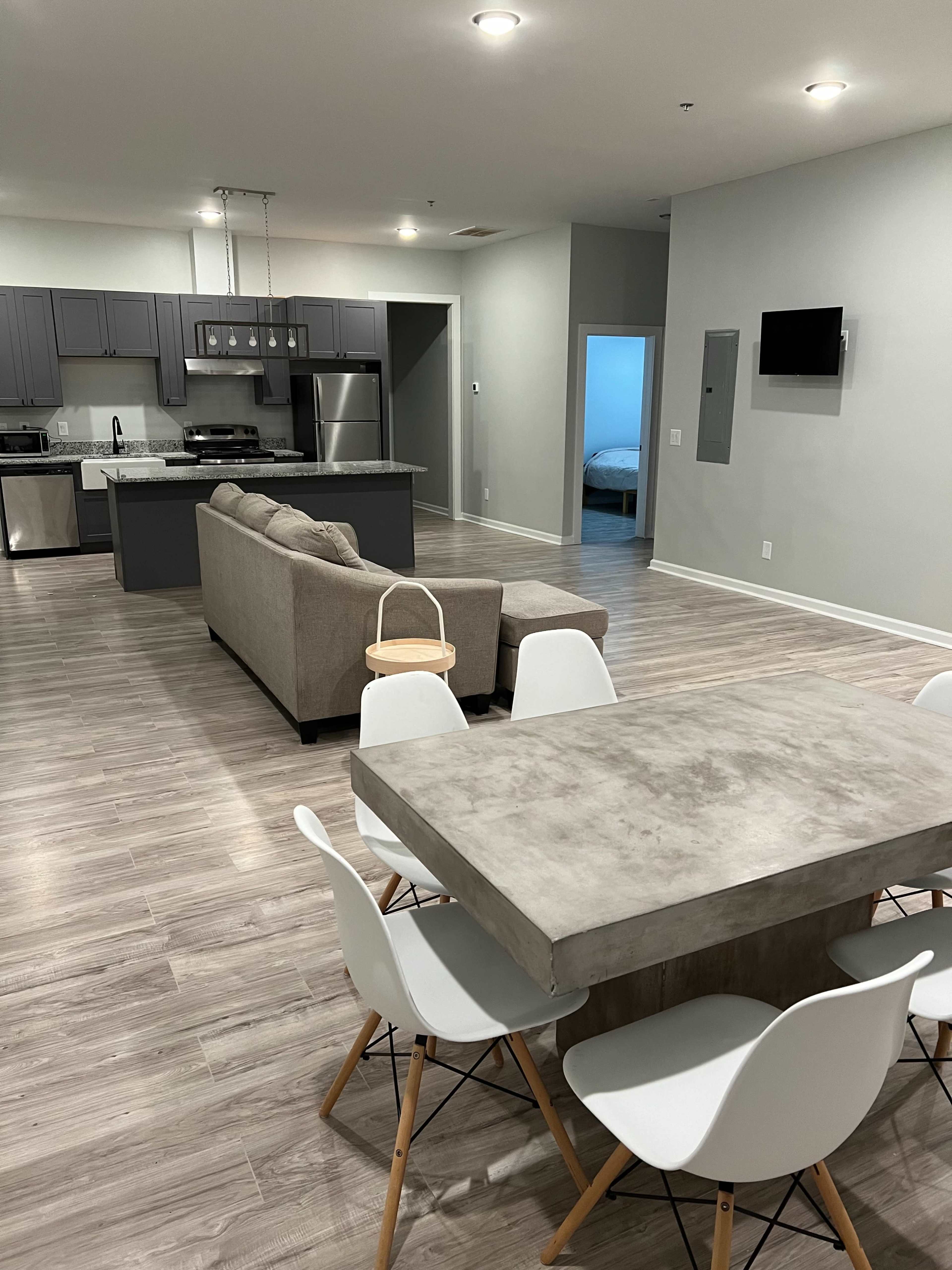 The image shows a modern open-concept living space with a kitchen, dining area featuring a concrete table and white chairs, and a living room with a gray couch.