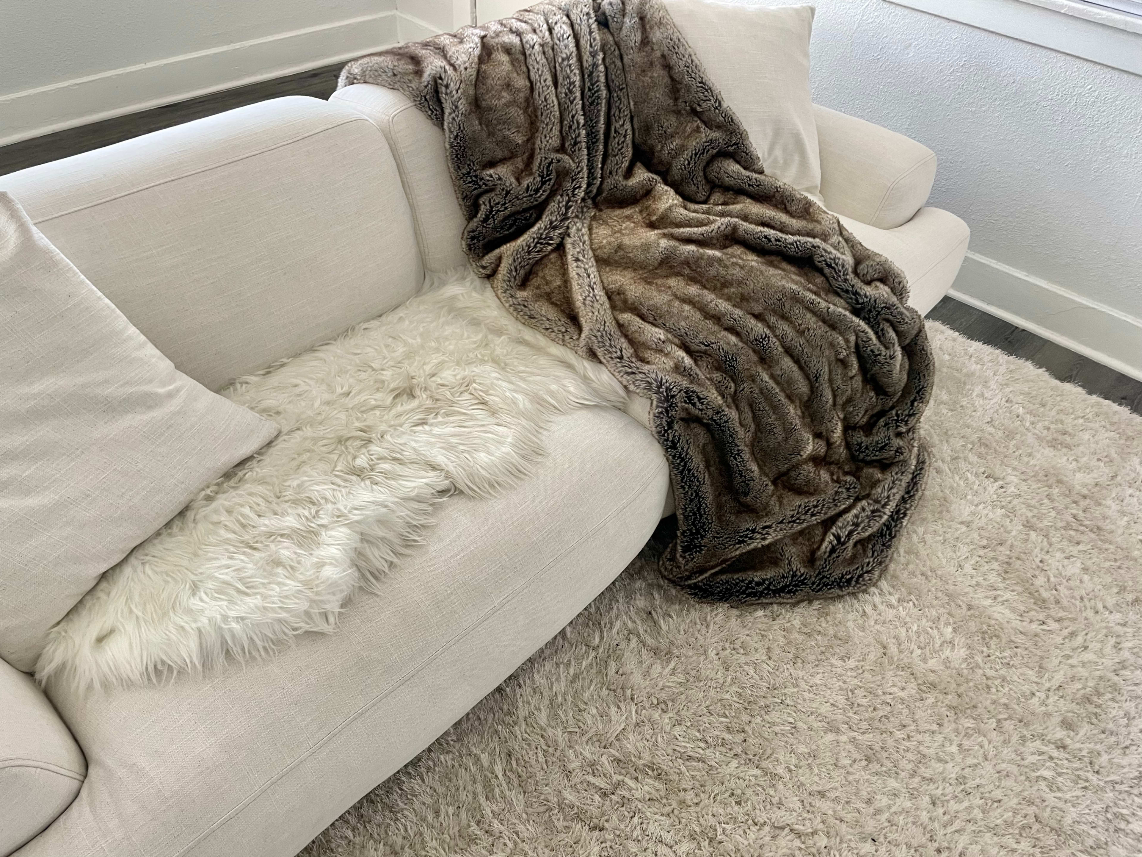 A light-colored sofa with two cushions features a soft, faux fur throw draped over one armrest, resting next to a plush area rug.