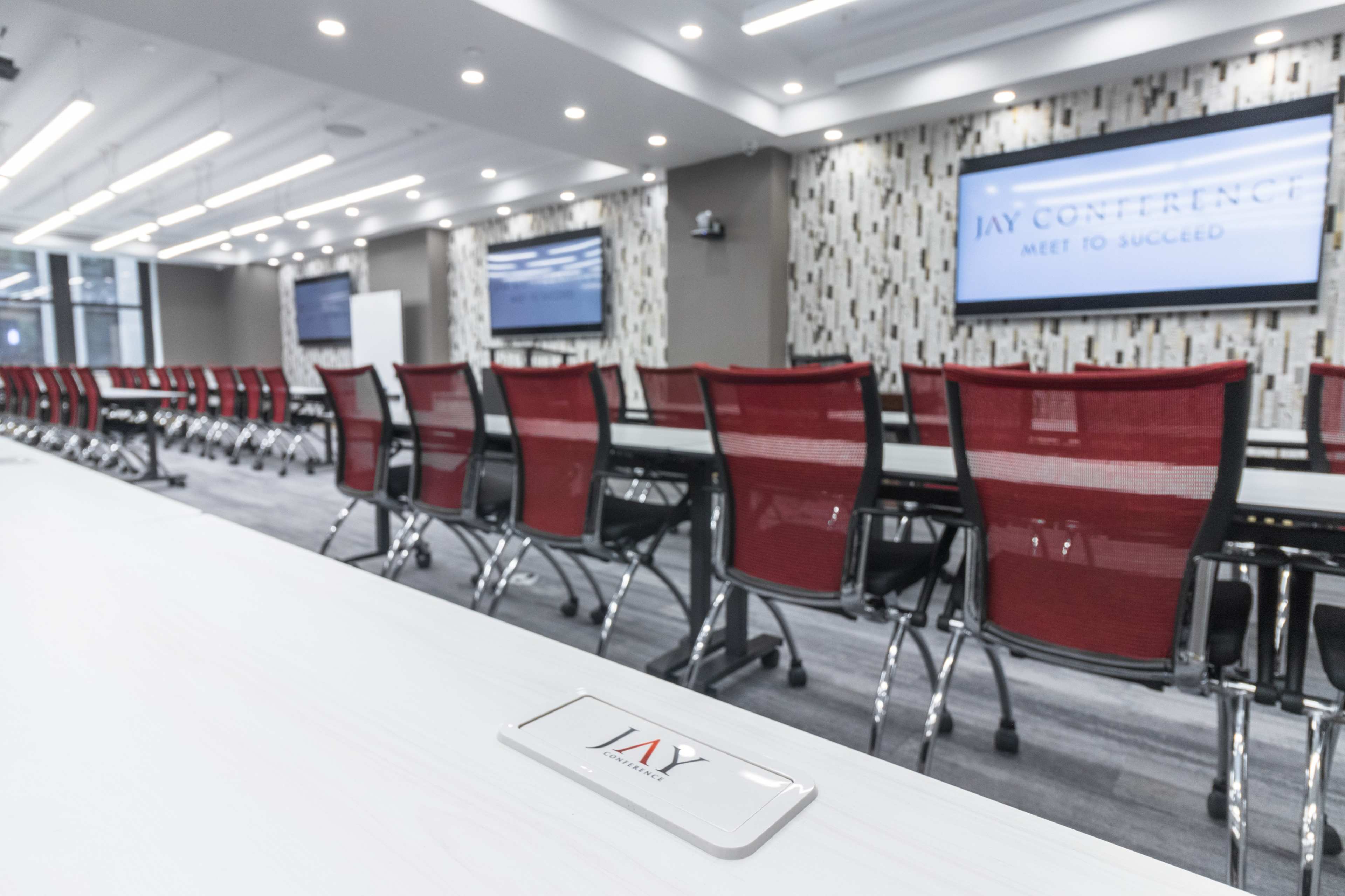 A modern conference room with rows of red and silver chairs arranged around white tables, featuring screens on the walls.