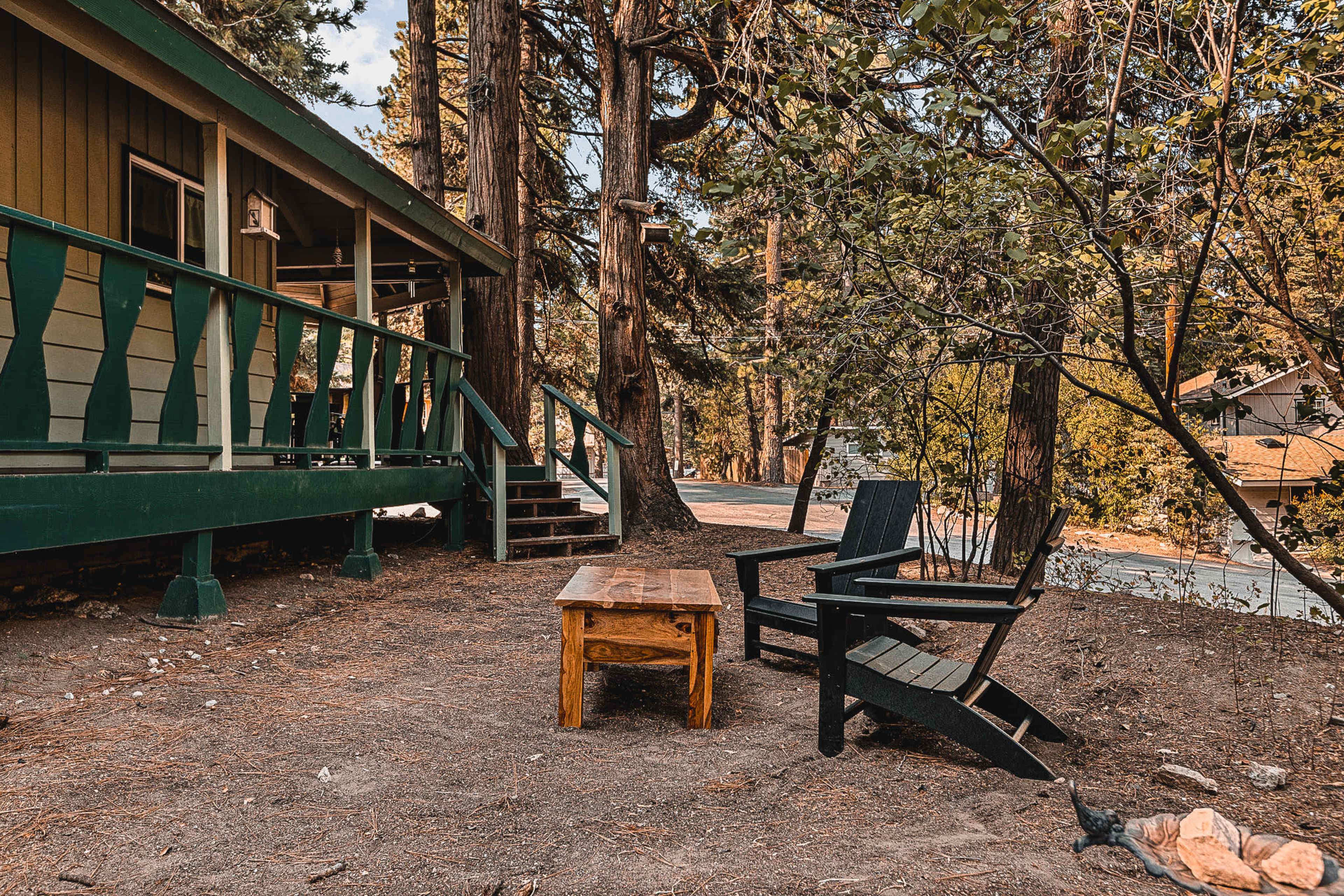 A wooden table sits between two black Adirondack chairs in a forested area next to a cabin.