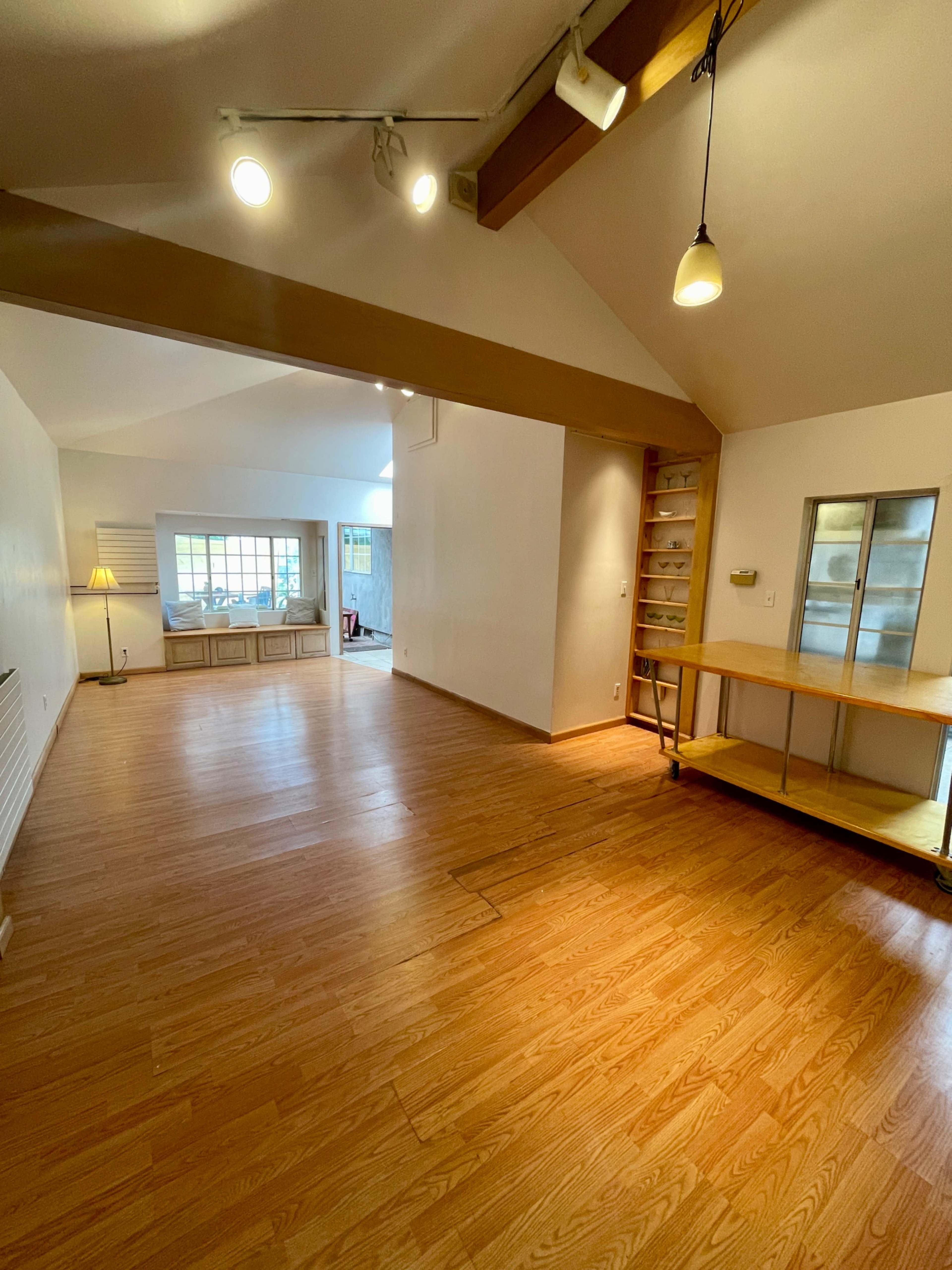 The image shows a spacious, well-lit room with wooden flooring, featuring a high ceiling and a small kitchenette area in the corner.