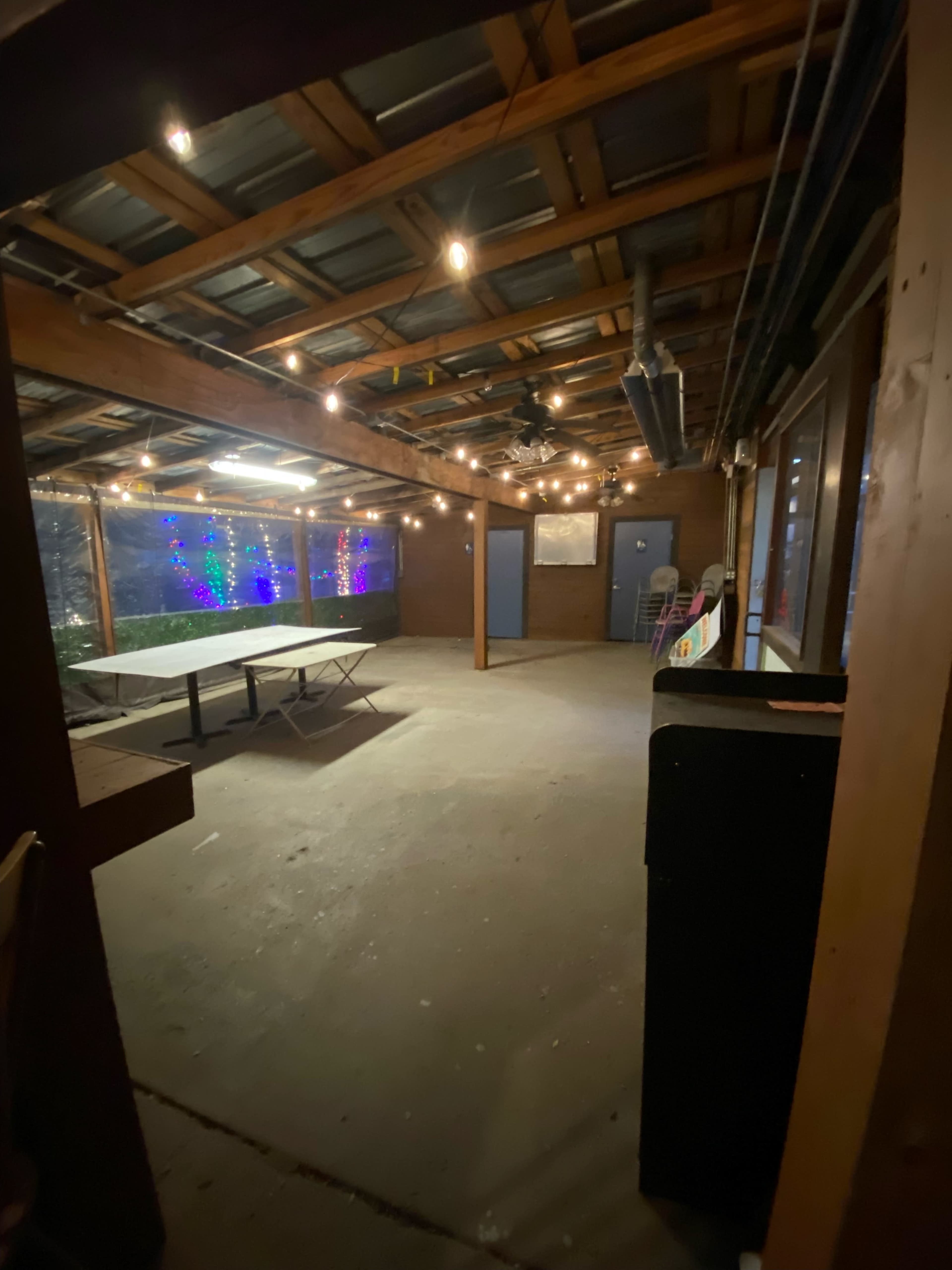An empty, covered outdoor space with a concrete floor, string lights overhead, and various furnishings along the walls.