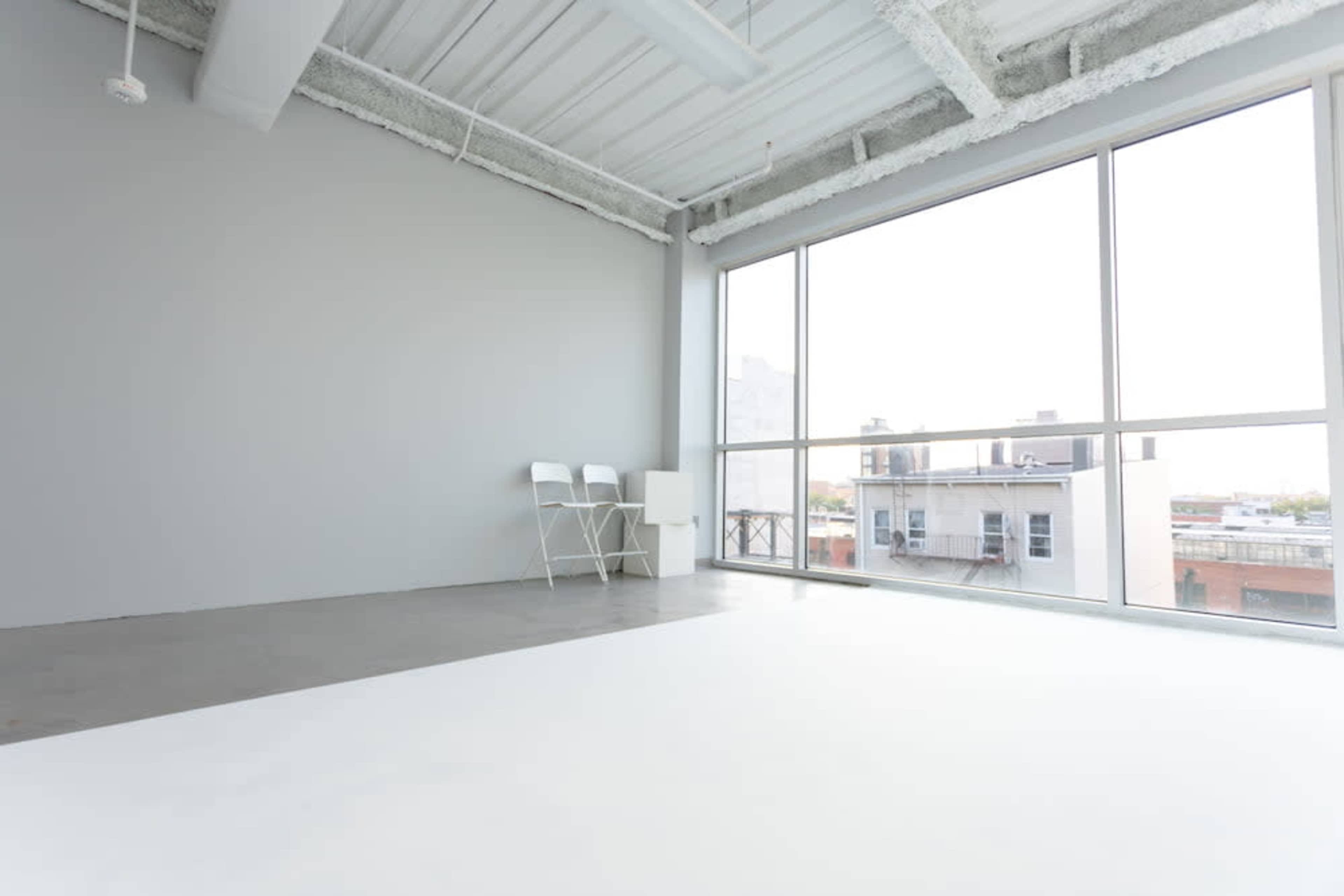 A spacious, minimalist interior with a large window overlooking a cityscape, featuring two chairs and a lamp against a light grey wall.